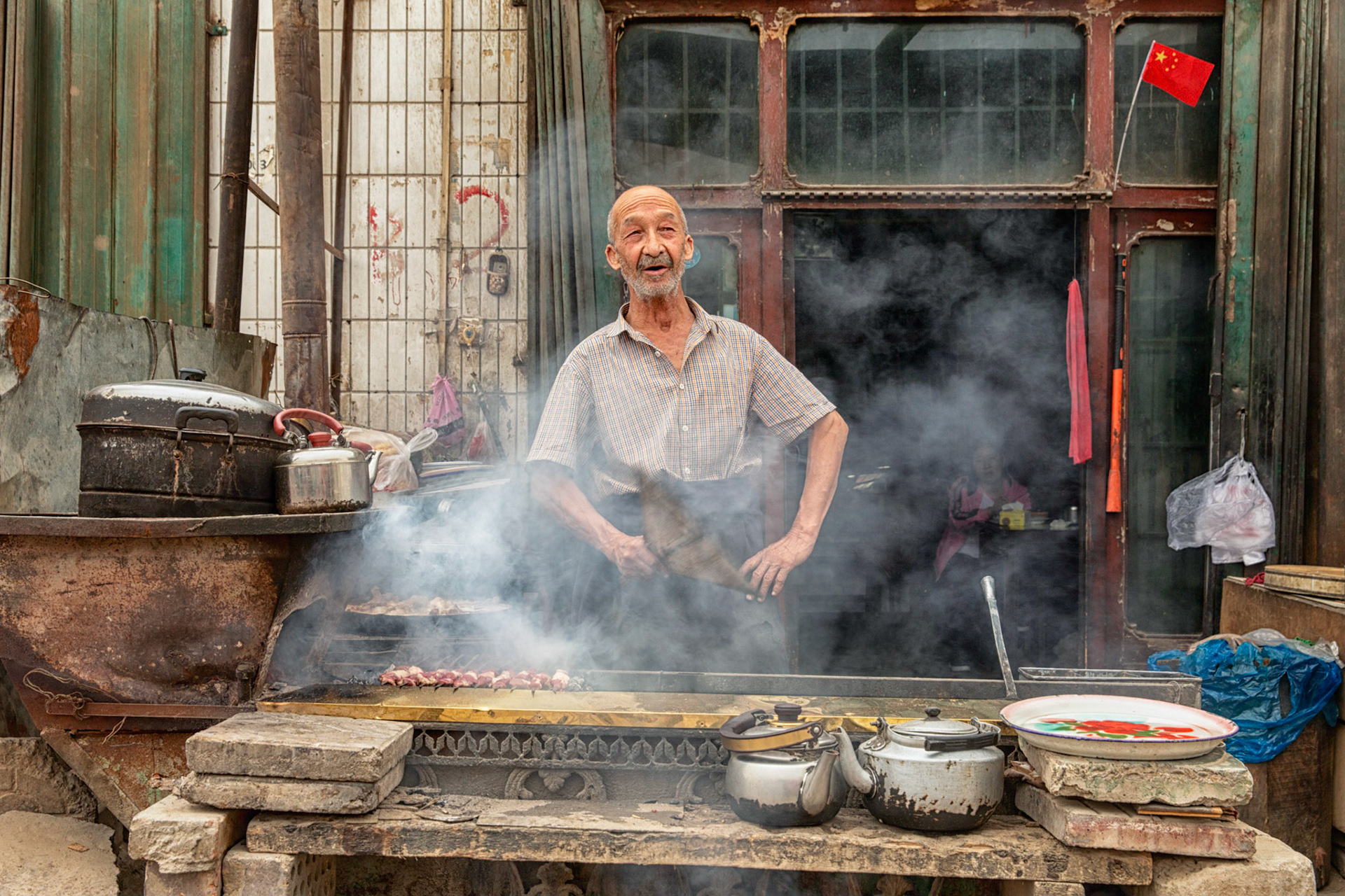 Hotan, China - September 13th 2018 :  Uyghur man, grillig kebap in his small streetside restaurant, Chinese flag in the window.