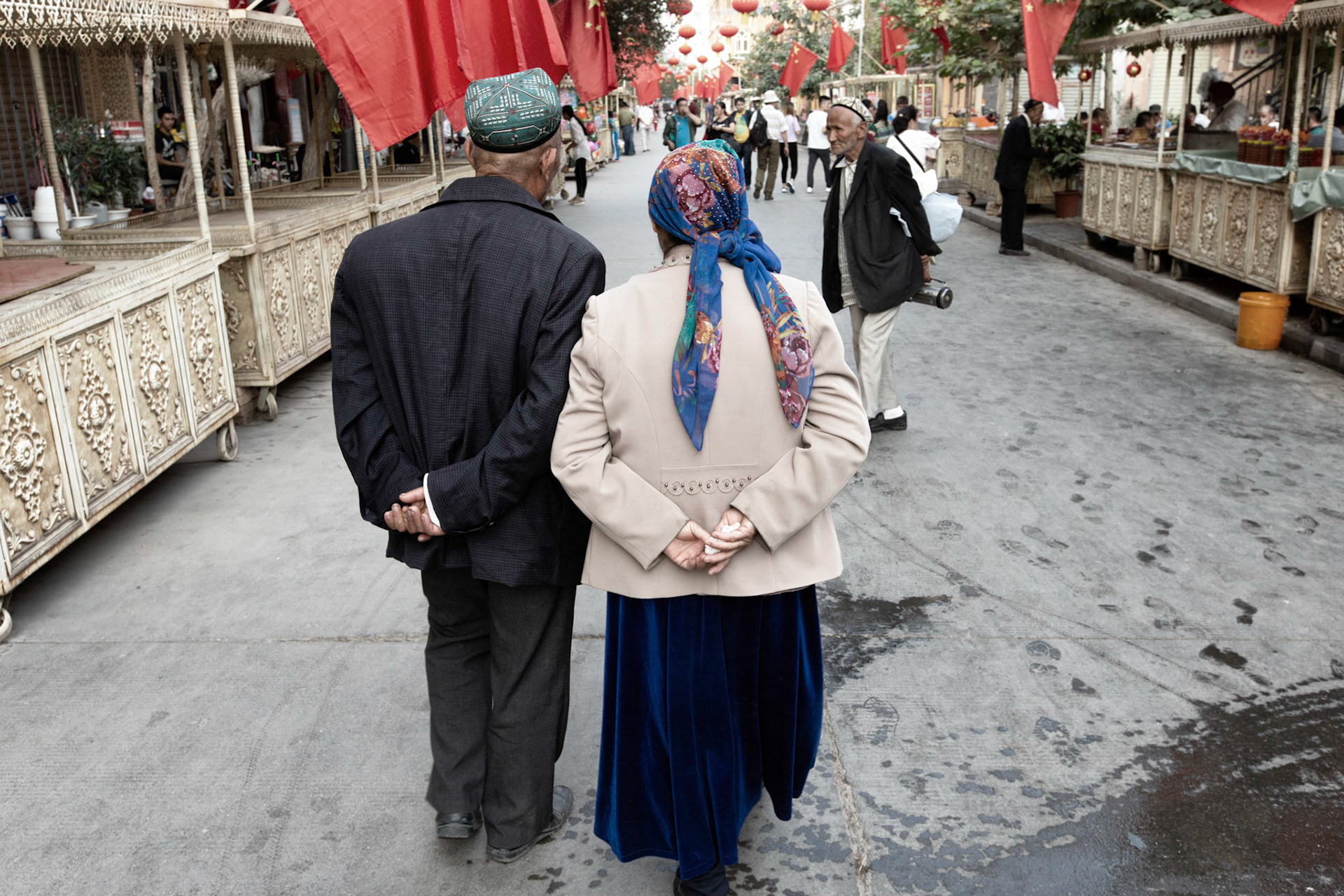 Kashgar,  China - September 11th, 2018 : Old age Uyghur couple walking together early evening on the Kashgar food market, which still hasn't fully opened yet. Chinese flags on each of the stalls.