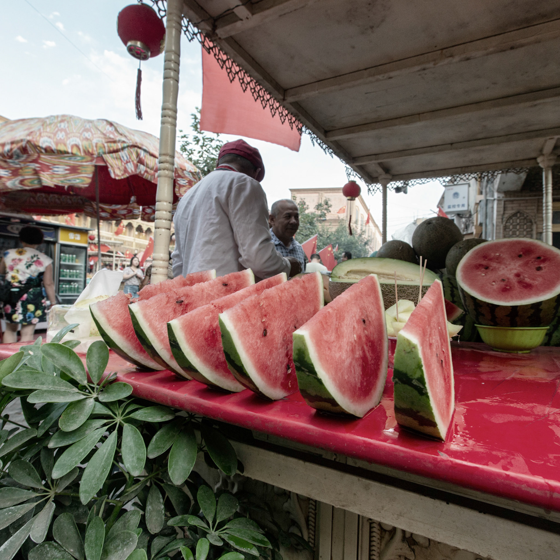 Kashgar,  China - September 11th, 2018 : Watermelon at a small food stall in the Kashgar food market, two men chatting with, people walking in the background