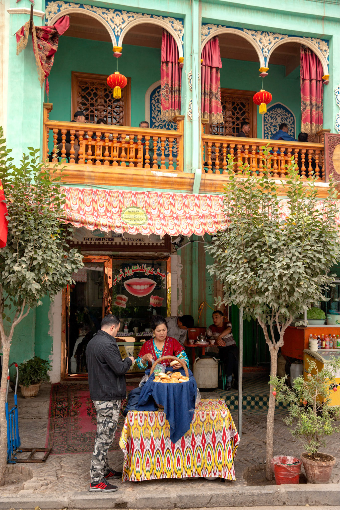 Kashgar, Xinjiang, China - September 5, 2018: Uyghur woman selling nan bread in front of an old tea house in Kashgar. Man standing and buying bread. People drinkin tea on the balcony.