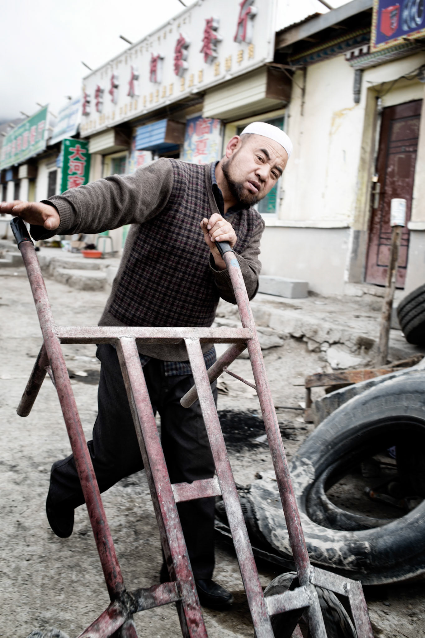 Qinghai, China - September 18th, 2015 : Muslim man wearing a hat typical for muslim, pushing a pushcard in a tyre shop in a roadside village in Qinghai, China