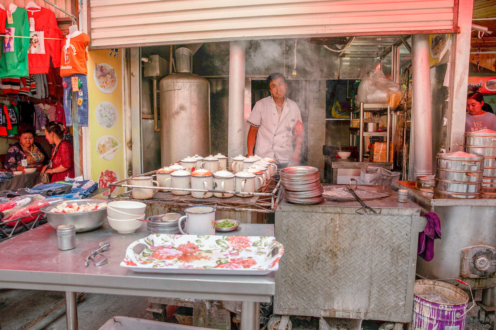 Kashgar, Xinjiang, China - September 5, 2018: Cook at a kitchen at a food stall in the Kashgar Grand Bazaar. females chatting in the background. Mugs for lamb meat soup and stacks of steamed bun pans visible