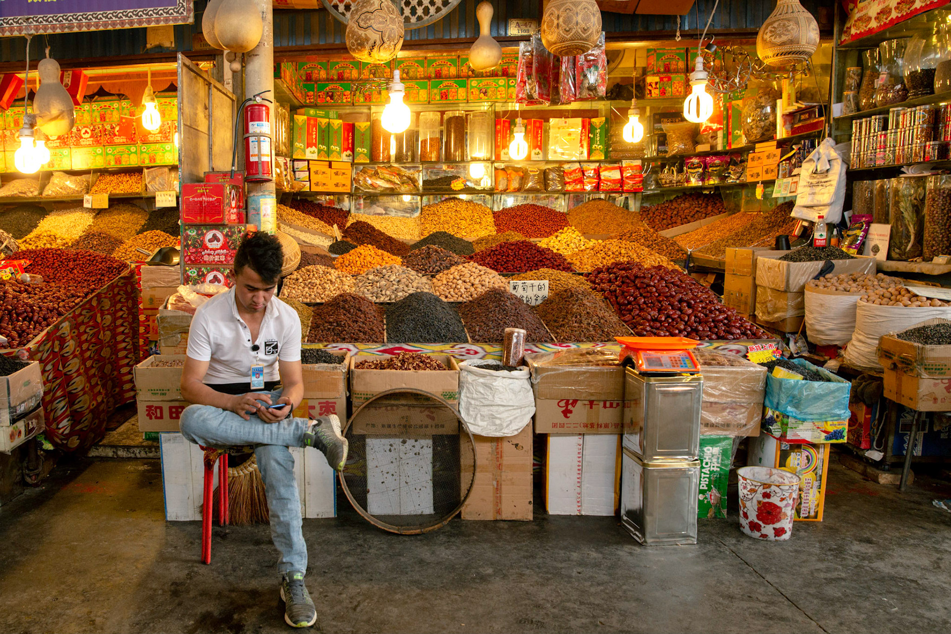 Kashgar, Xinjiang, China - September 5, 2018: Shopkeeper sitting in front of his shop in the Kashgar Bazaar, checking his mobile phone. Many spices, nuts, tea and other produce in the background