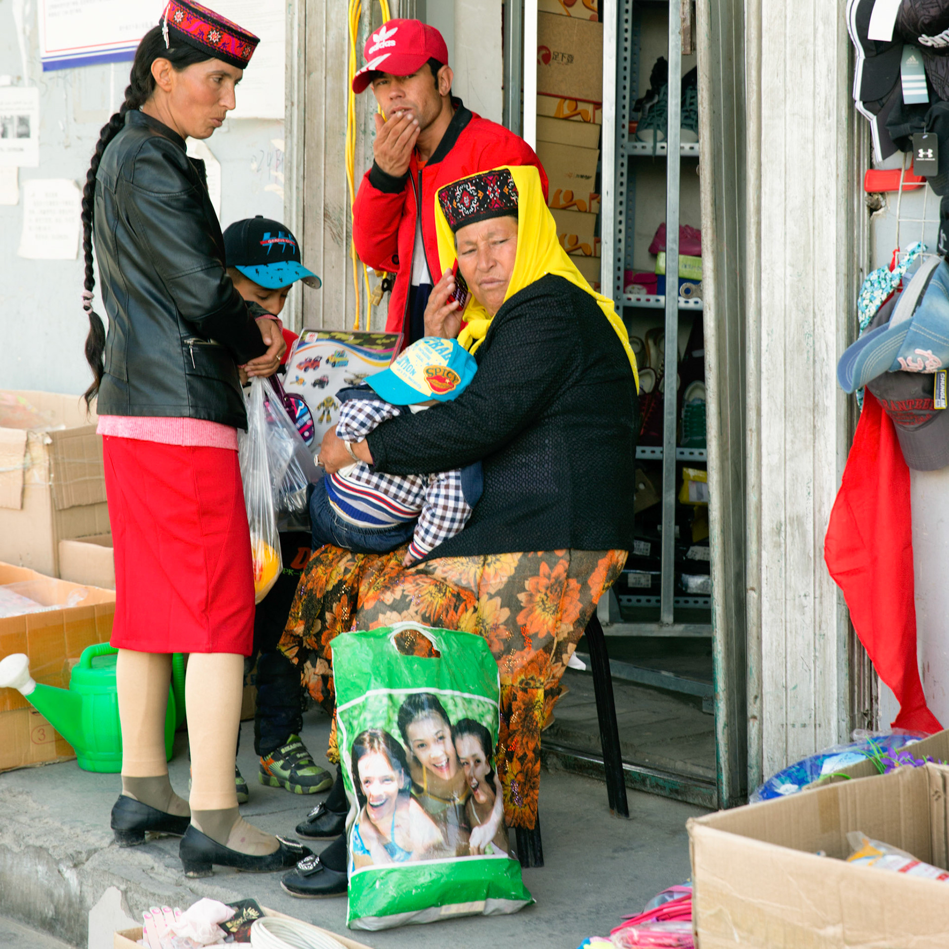 Tashgurkan, China - September 7th, 2018 : Group of women wearing typical tajic dress and hat sitting and standing in front of a store.