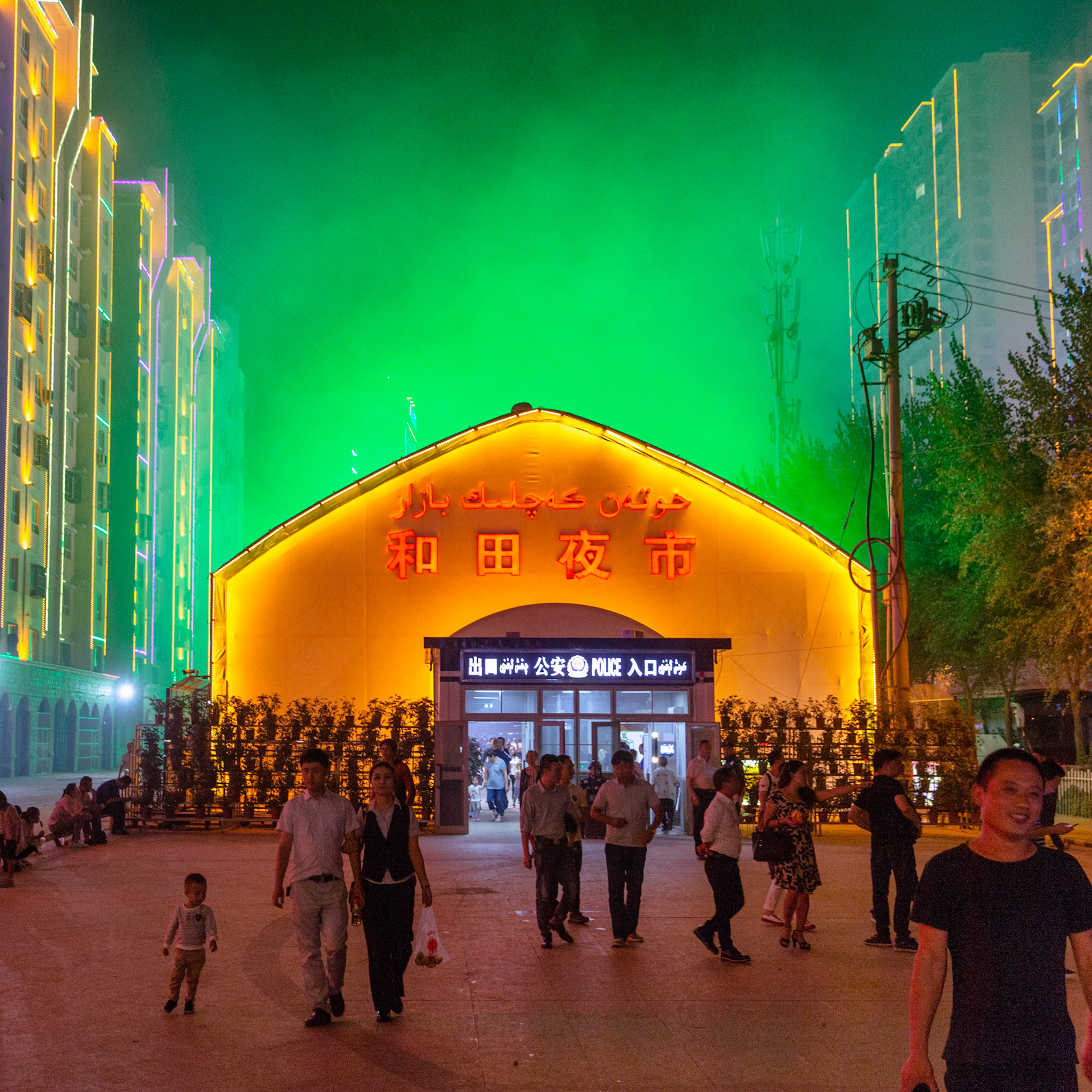 Hotan, China - September 12th 2018 :  Outside view of the Hotan Night market, sign on the building says "Hotan Night market in both Uyghur and Chinese,  Police checkpoint at the door and people walking in and out.