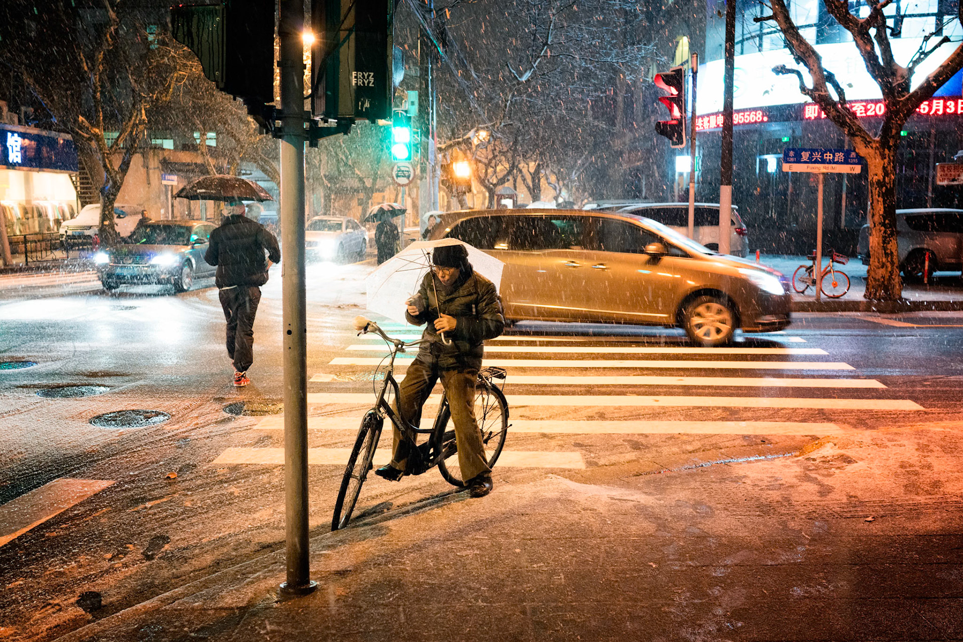 Shanghai, China - January 25th 2018 : Evening image of a man with a bicycle holding a white umbrella and standing still at a zebra crossing drinking something. Cars and people in the background