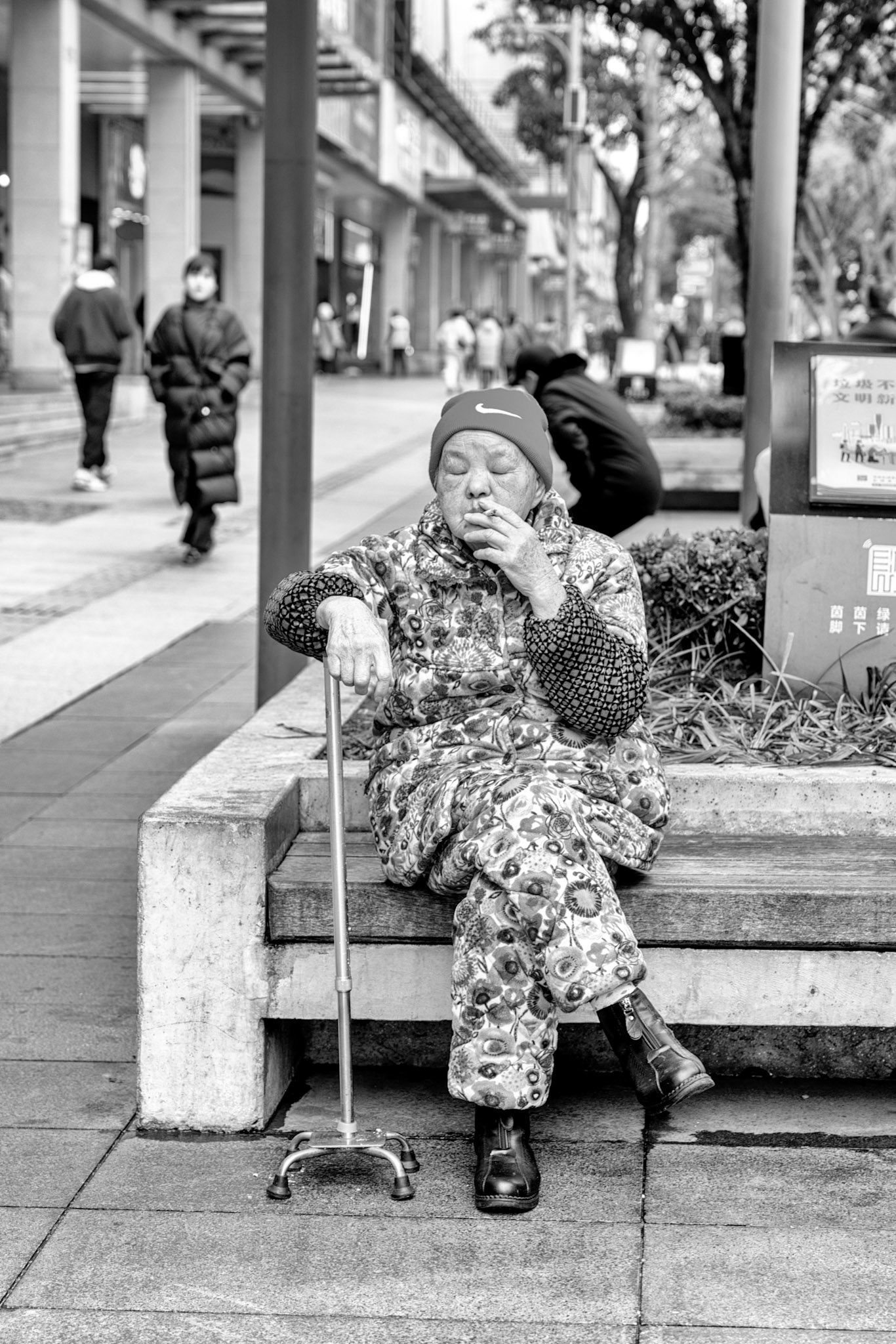 Changsha, China - 27 Feb 2024: An elderly resident, wearing padded clothing, takes a moment to rest on a bench along a busy street in Changsha.