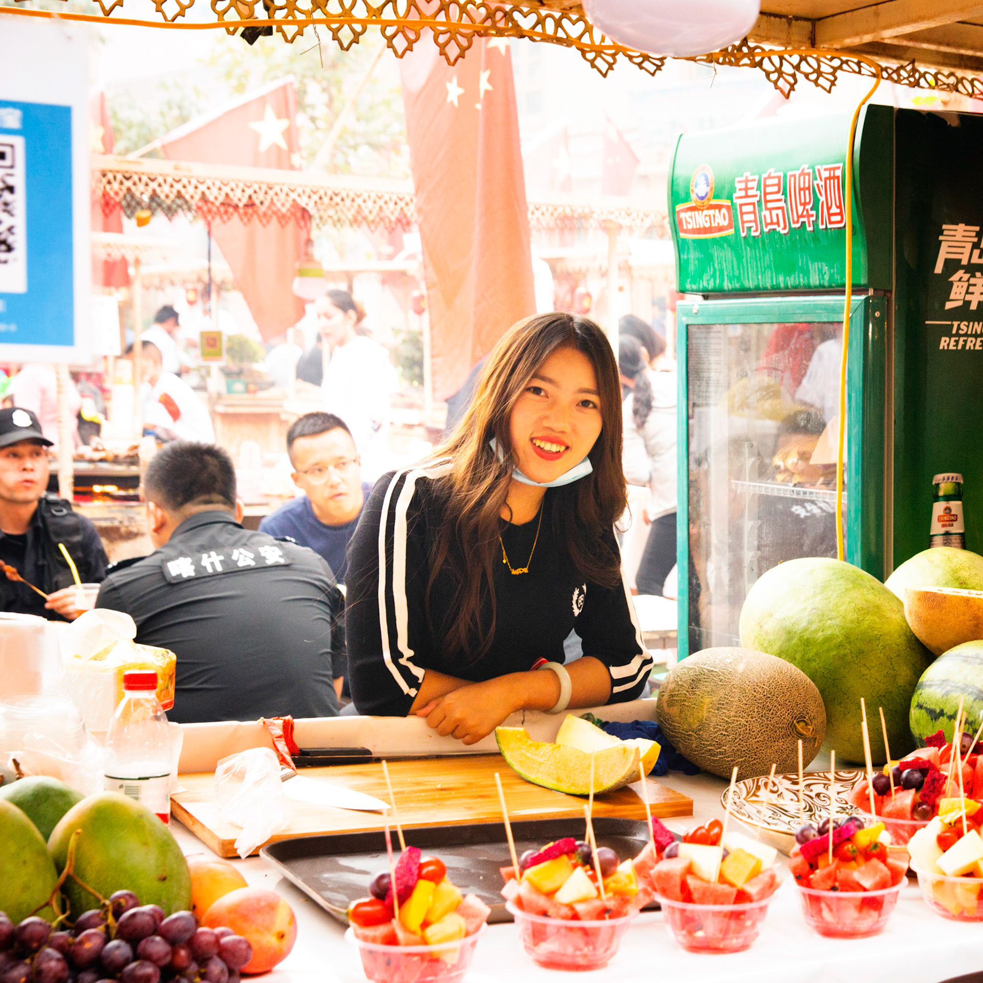 kashgar, Xinjiang, China - September 5, 2018: Chinese girl selling fresh fruit at the Kashgar food market, police sitting at a table eating in the background