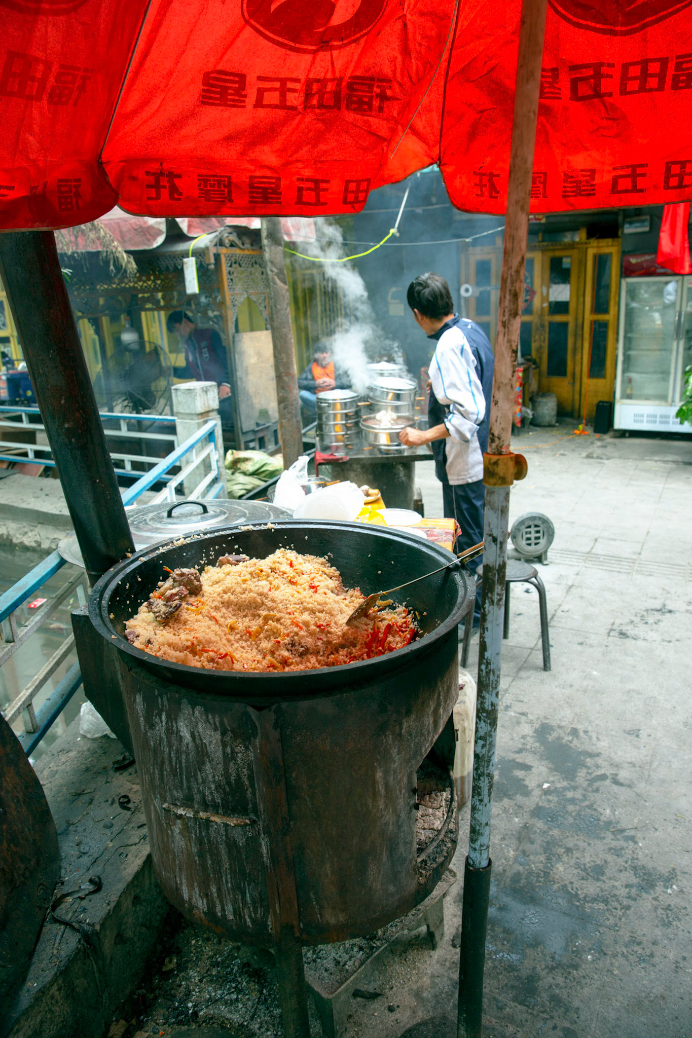 Tashgurkan, China - September 7th, 2018 : Rice and lamb meat kooking on a coal stove, steamy buns being prepared in the background.