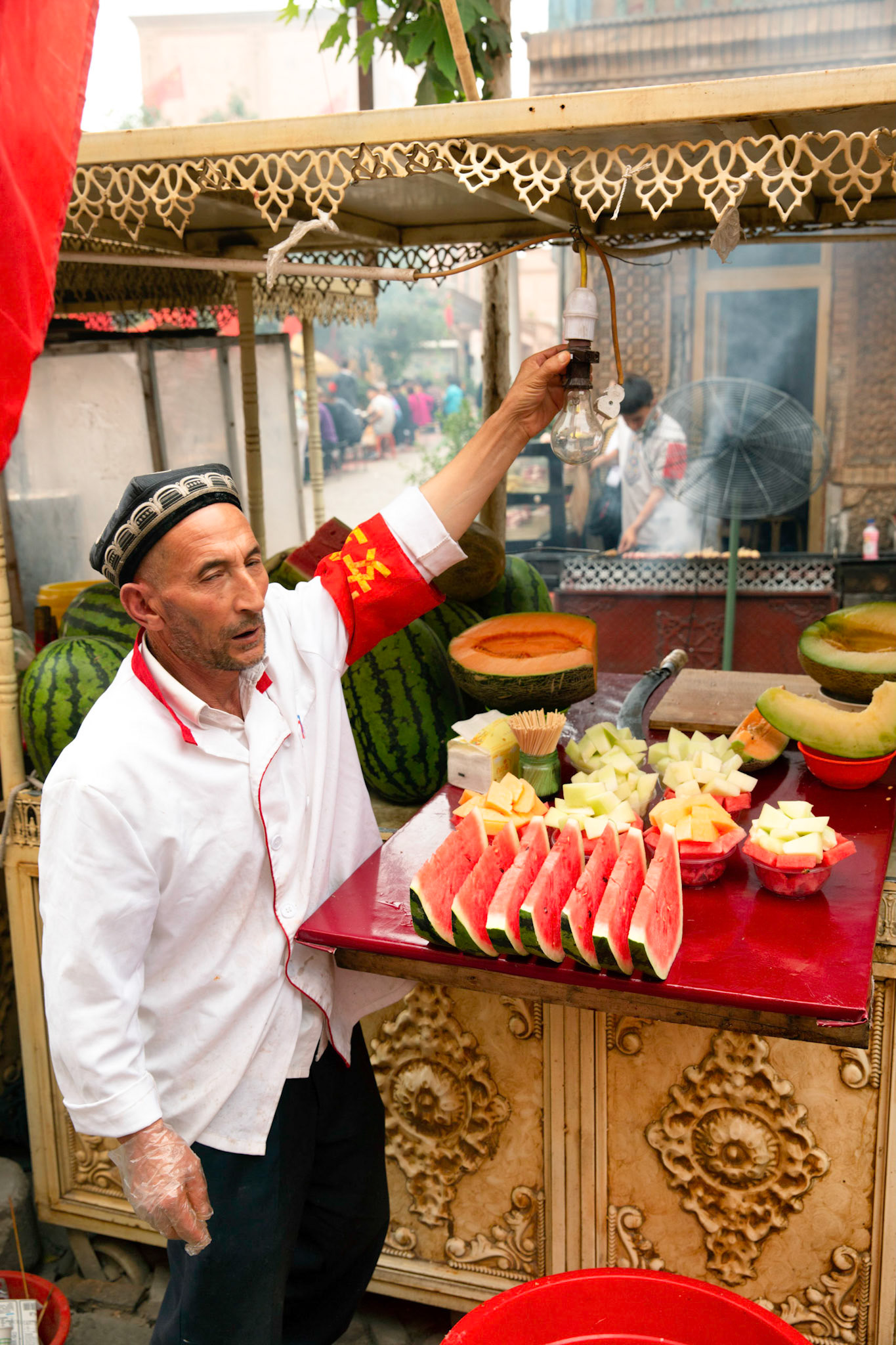 Kashgar, Xinjiang, China - September 5, 2018: Man in selling fresh fruit in a a fruit stall at the Kashgar night market.Grill and smoke in the background, people eating in the background