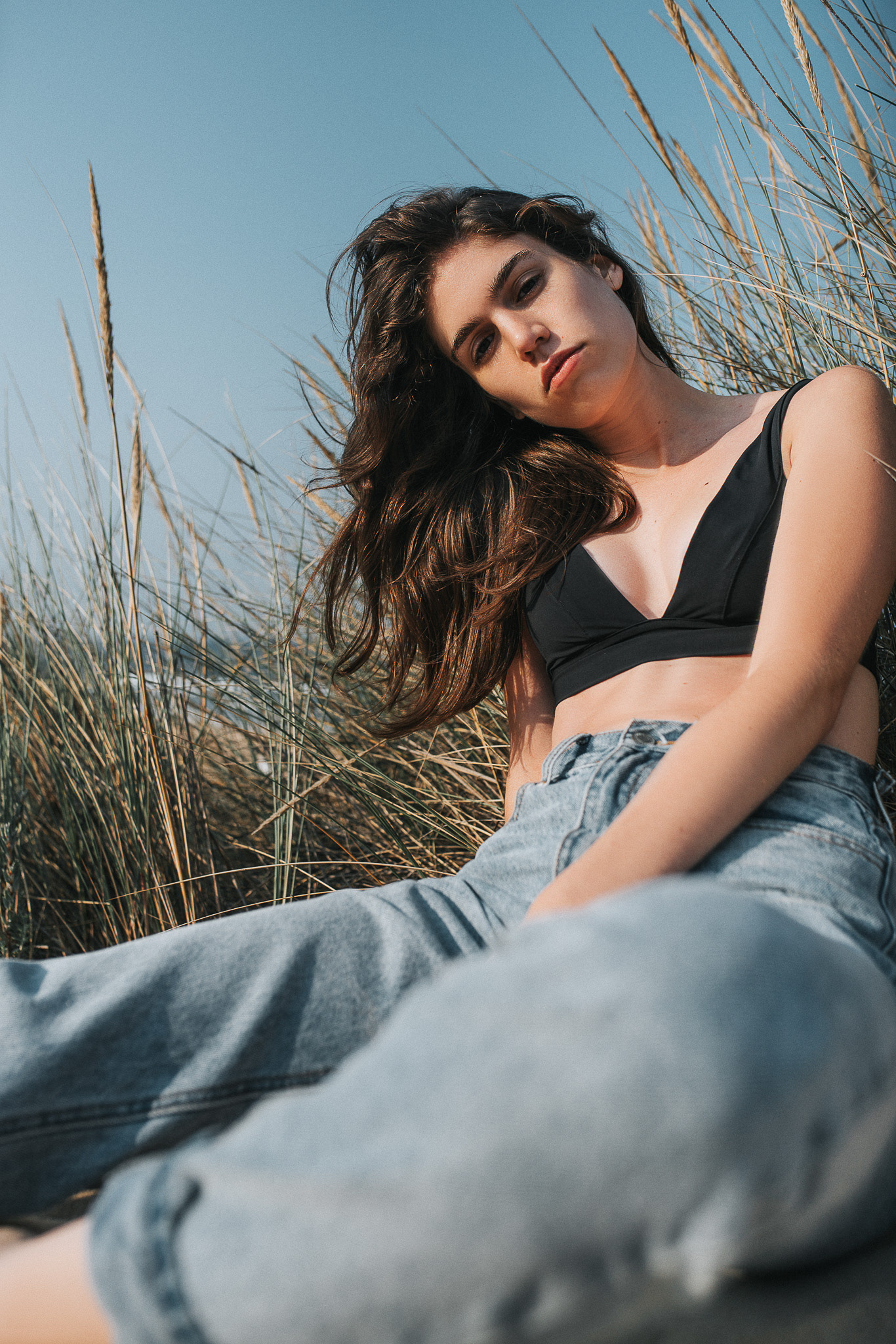 Strandshooting mit Helena – elegante Pose im Sand mit sanften Wellen im Hintergrund.