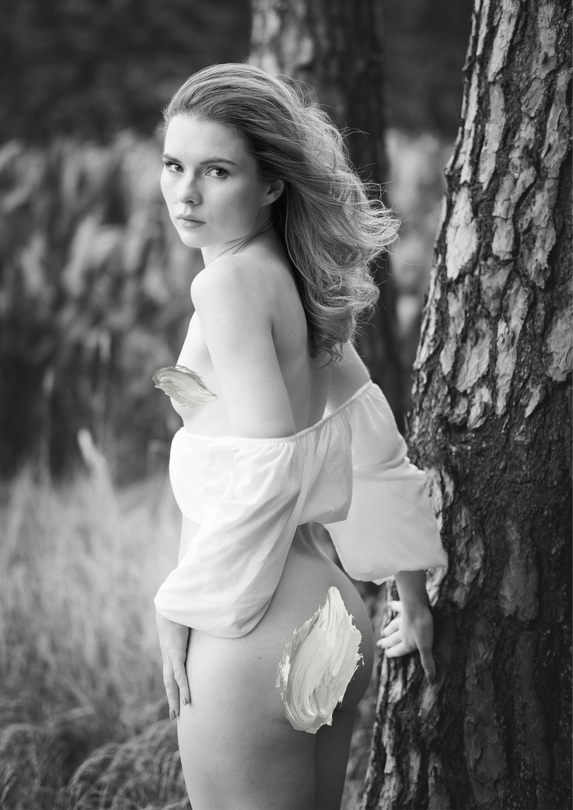 Fine art black-and-white portrait of a woman with long flowing hair standing beside a pine tree in a forest, wearing an off-shoulder white blouse and looking directly at the camera in a moody outdoor setting.