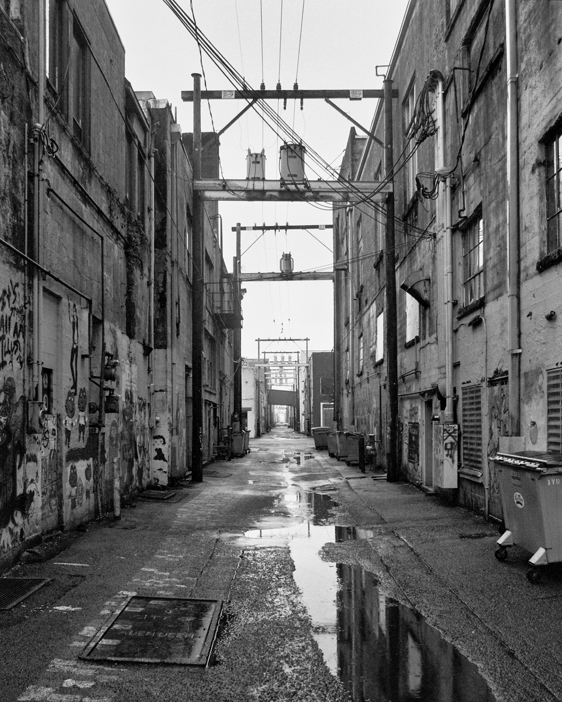 Black and white photo of a claustrophobic alleyway in Aberdeen, WA. Graffiti covers the walls and the sky reflects in puddles on the concrete.
