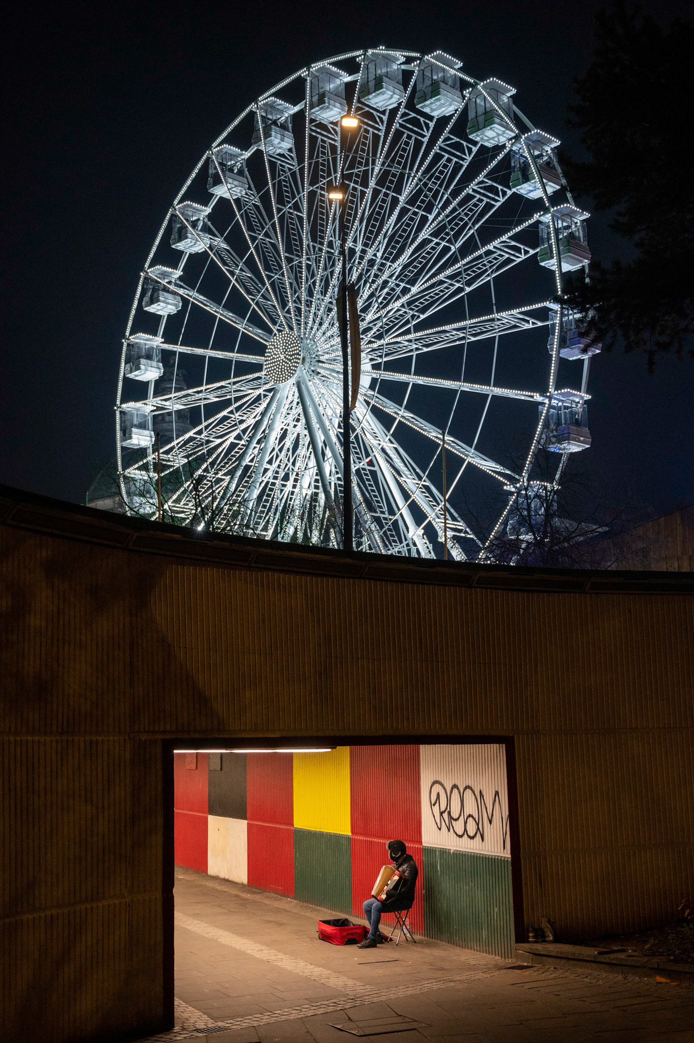 Two sides to a city. Homelessness alongside affluence, a common site in most UK cities these days. Here in Cardiff the homeless issue seems worse than ever with evidence all around the city centre.In this picture on a freezing cold December night in the shadow of the Ferris wheel at the festive Winter Wonderland and only a stones throw from City Hall a homeless person sits playing for passing revellers.