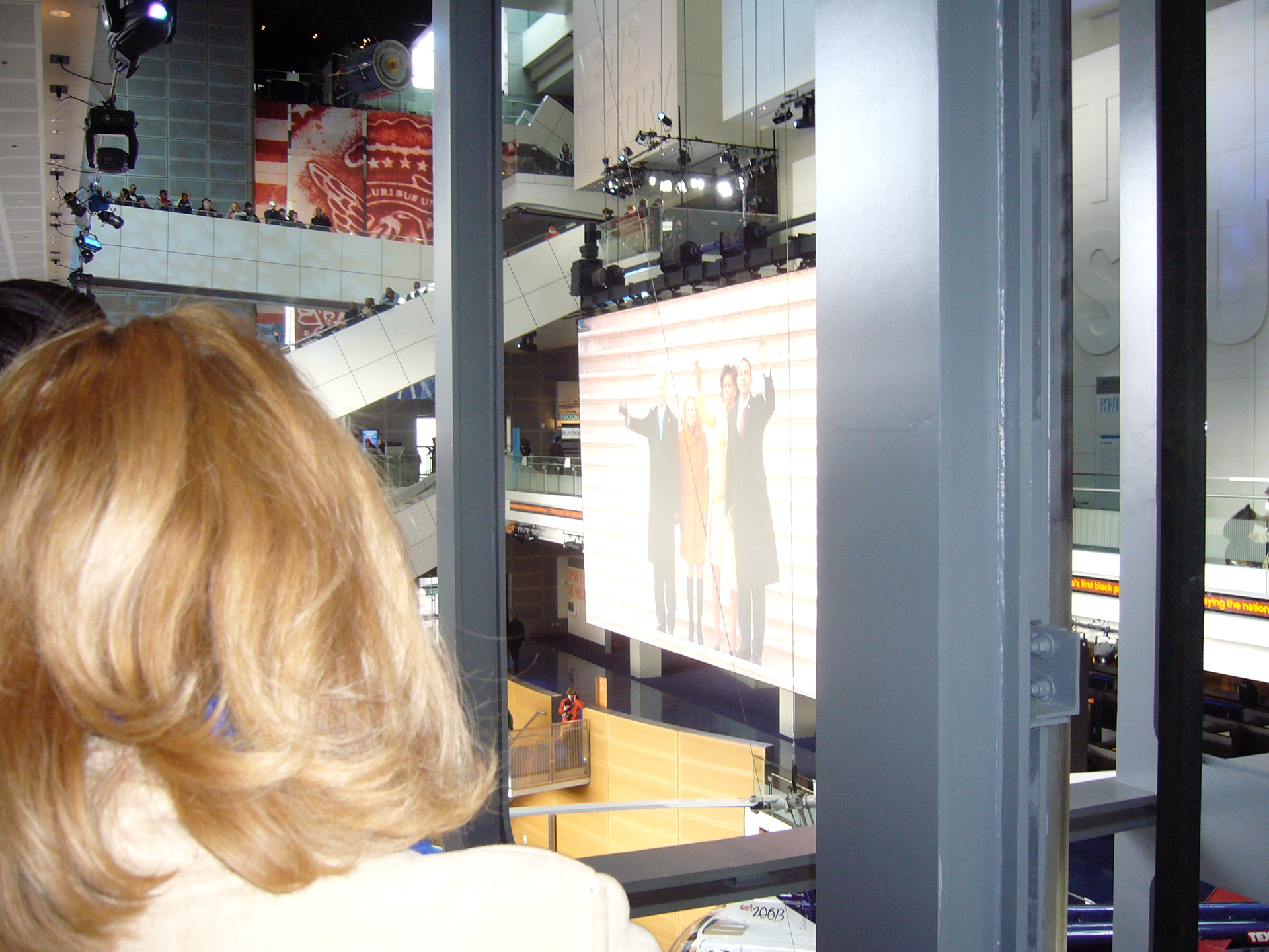 At top right news anchors cover the event while below a large screen shows the Obamas and Biden's greeting the crowd