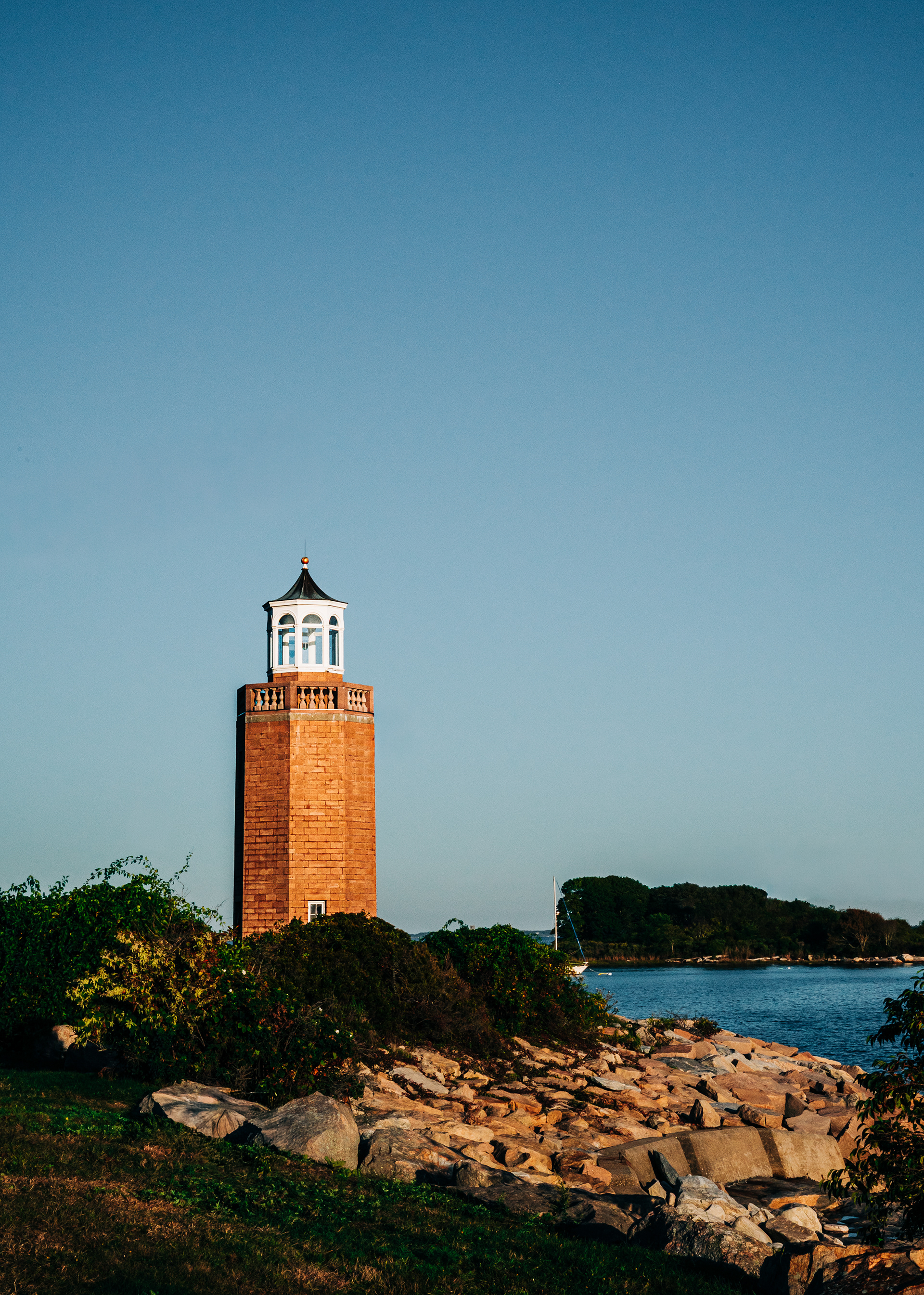   A tall lighthouse made of stone, standing on a rocky shore by the water, under a clear blue sky. 
