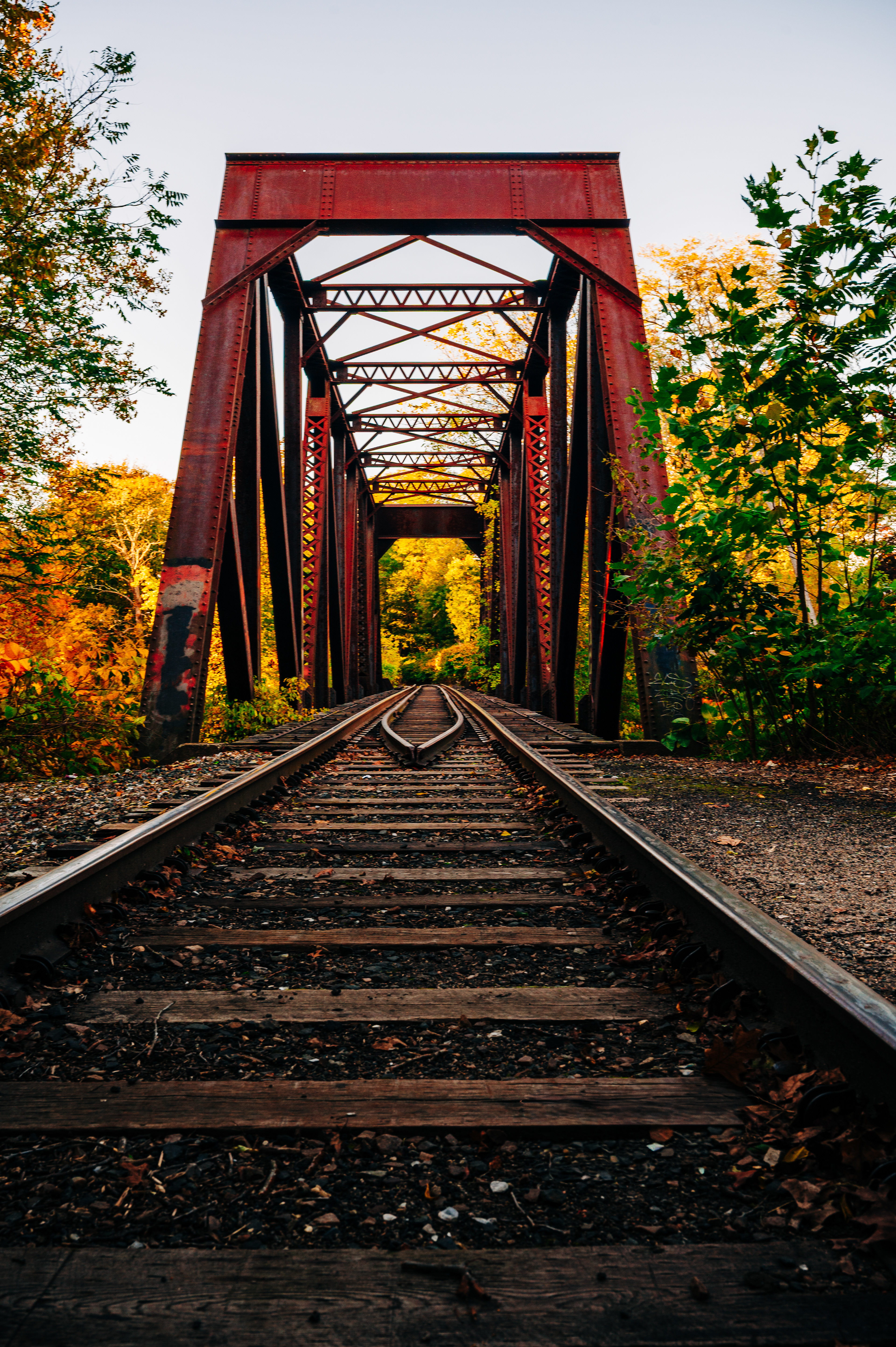   A red metal train bridge with green trees and tracks in the foreground. 