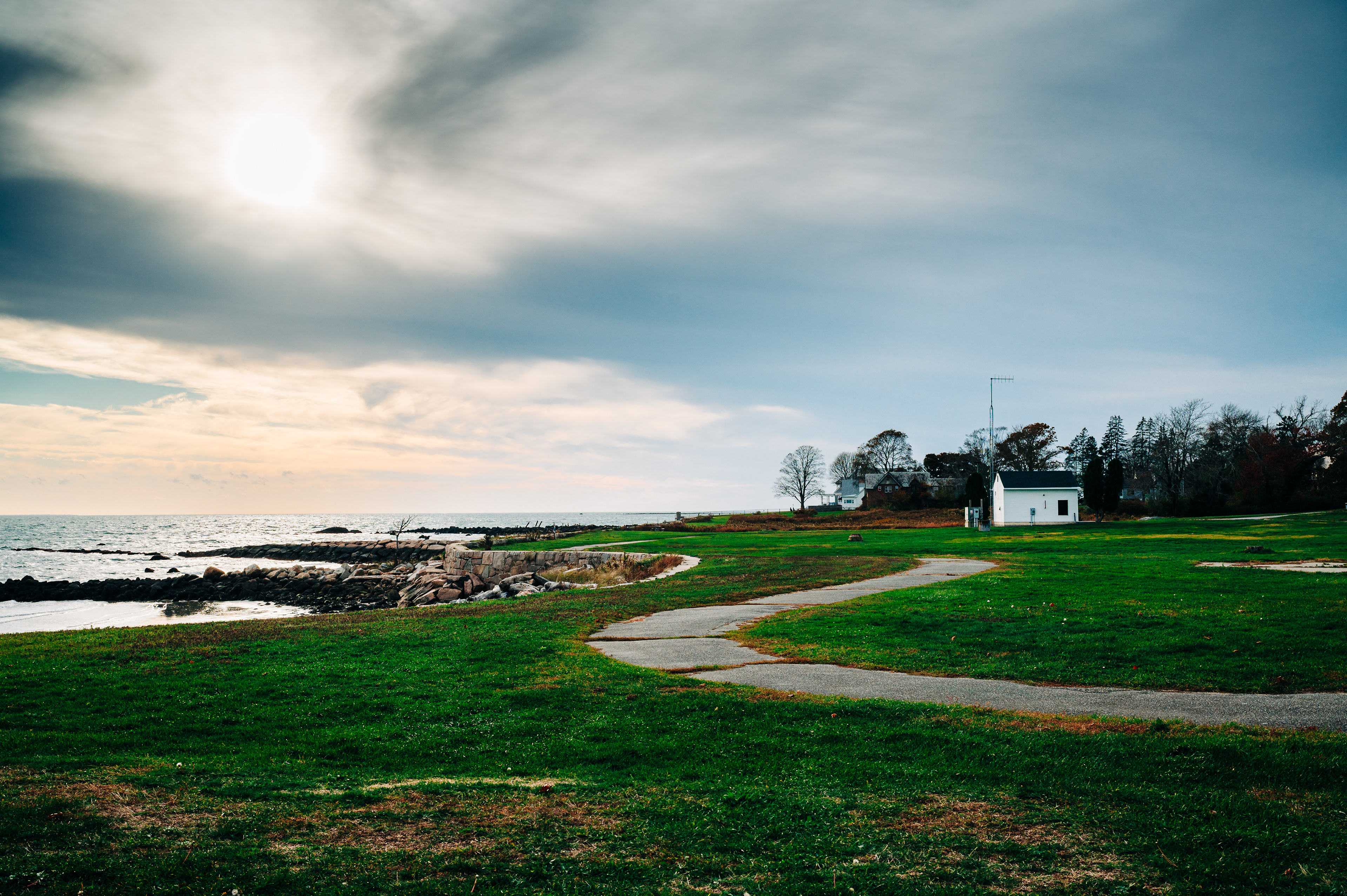   A coastal view with a winding path, grass, and a small white building near a rocky shore under a cloudy sky. 