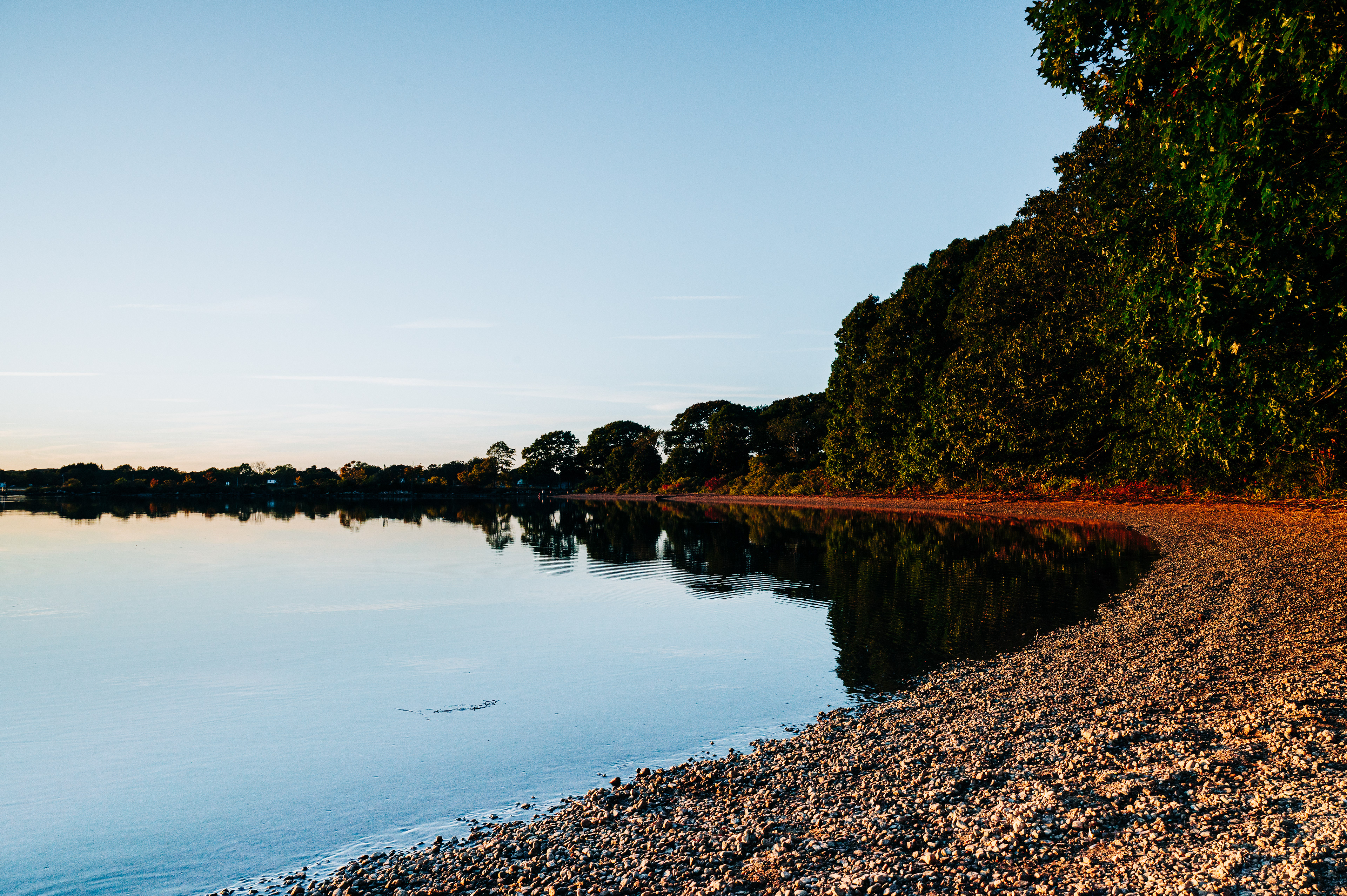   Calm lake reflecting trees and the sky, with a pebbly shore in the foreground. 