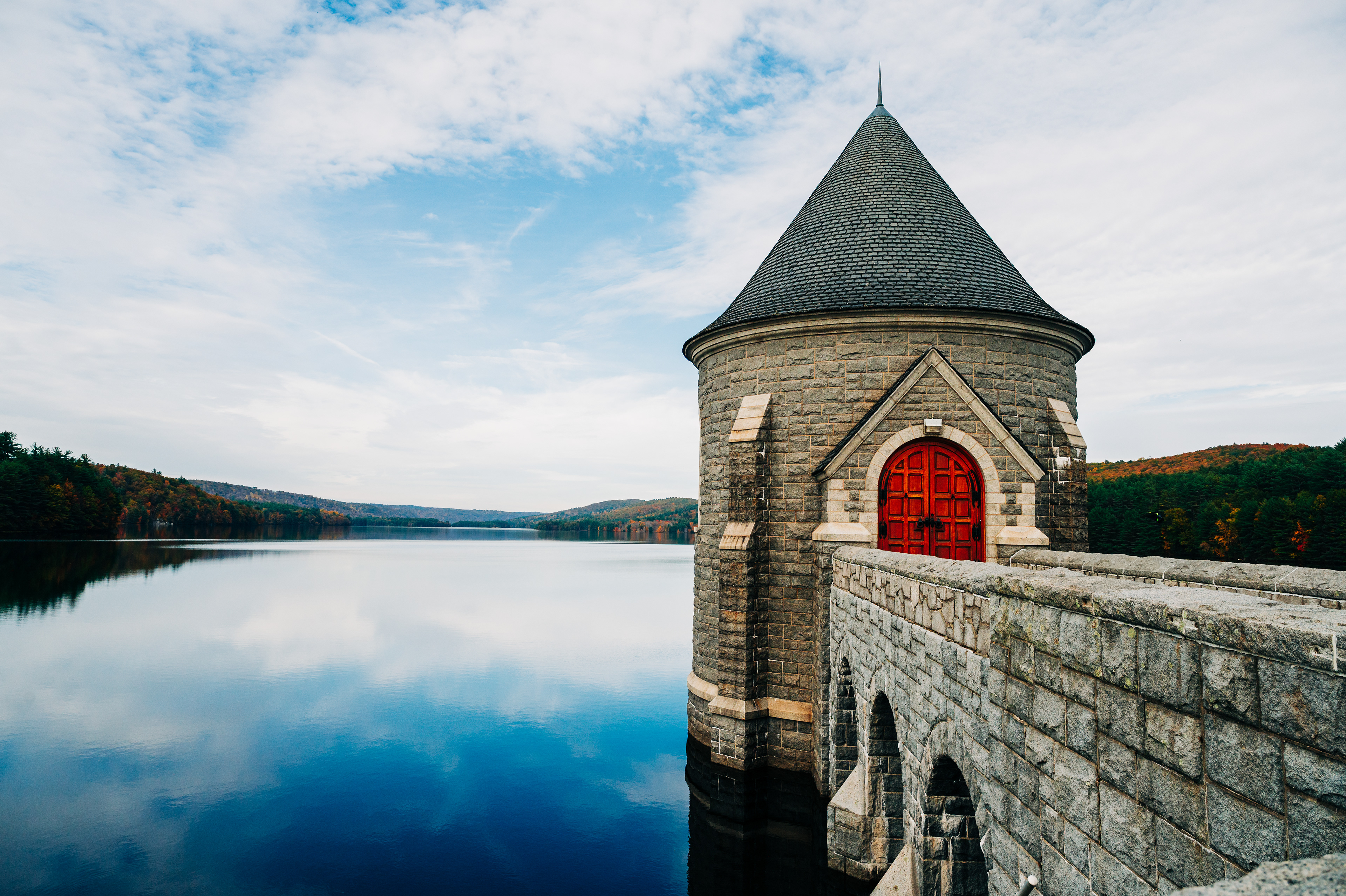   A stone tower with a red door by a calm lake, surrounded by trees and a blue sky. 