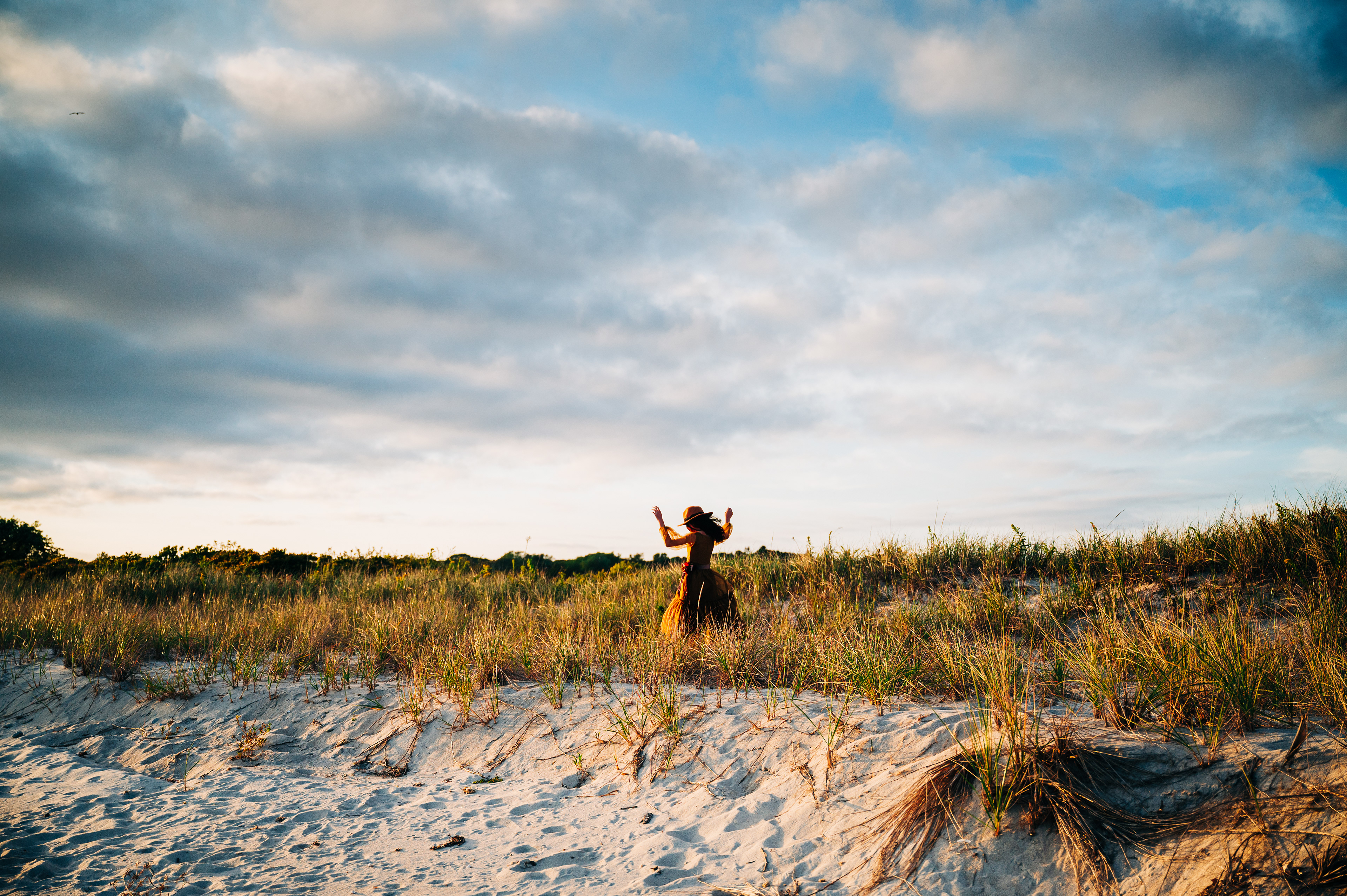   A person in a flowing brown dress dances joyfully on a sandy beach with tall grass. 