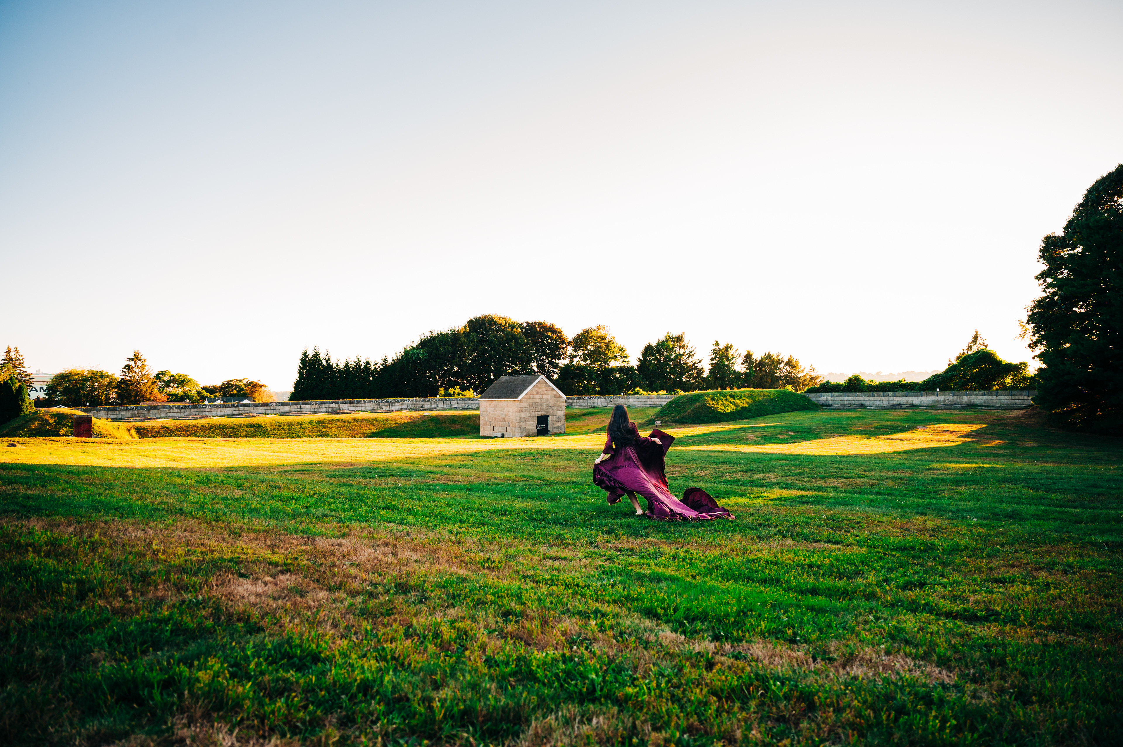   A person in a long purple dress stands in a grassy field, with a small building in the background. 