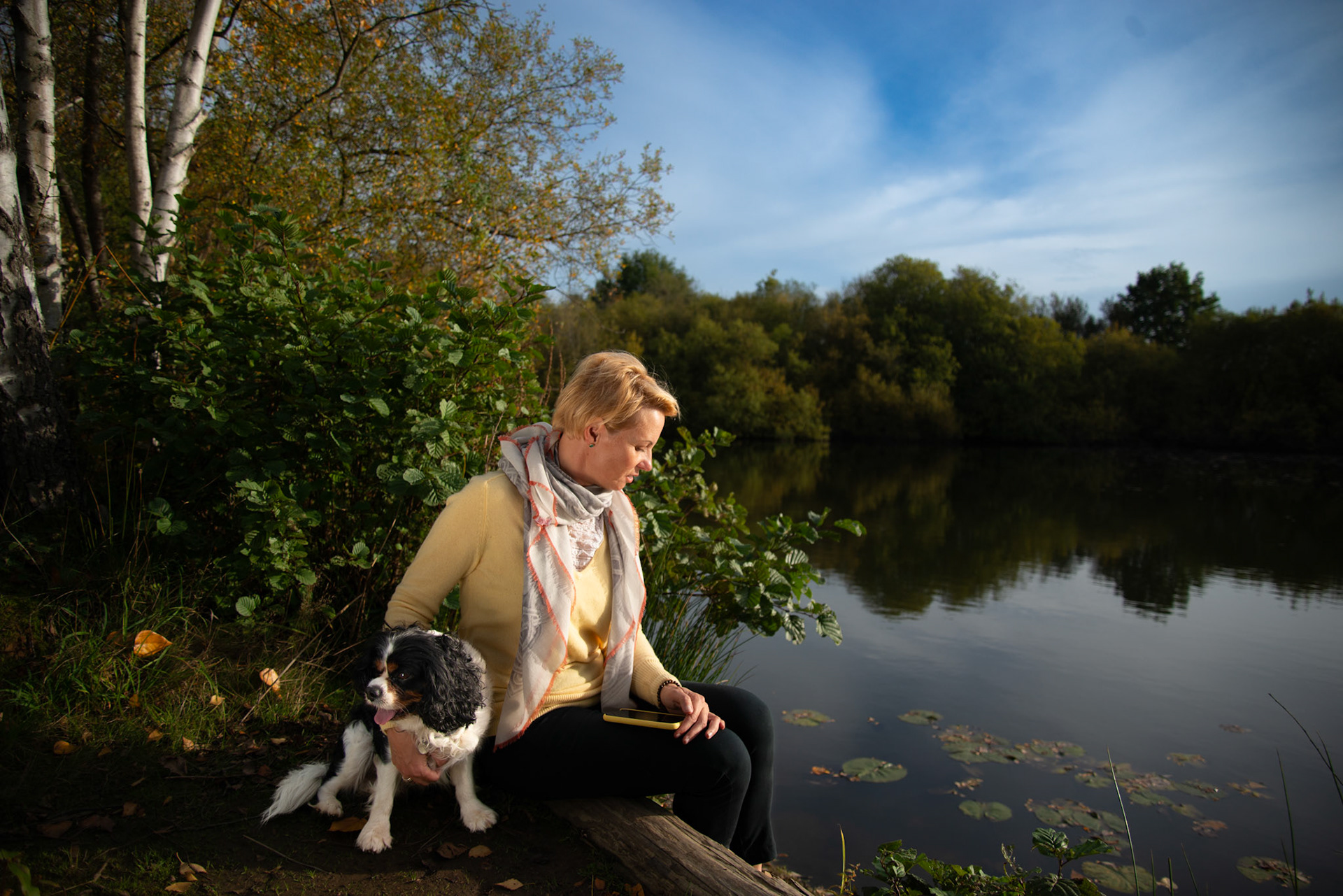 A woman in a yellow sweater and black pants sits contemplatively beside a small, fluffy black and white dog on a fallen log by a tranquil lake. The scene is peaceful, with autumn leaves scattered on the ground, water lilies floating on the water's surface, and a backdrop of dense trees reflecting the early signs of fall colors