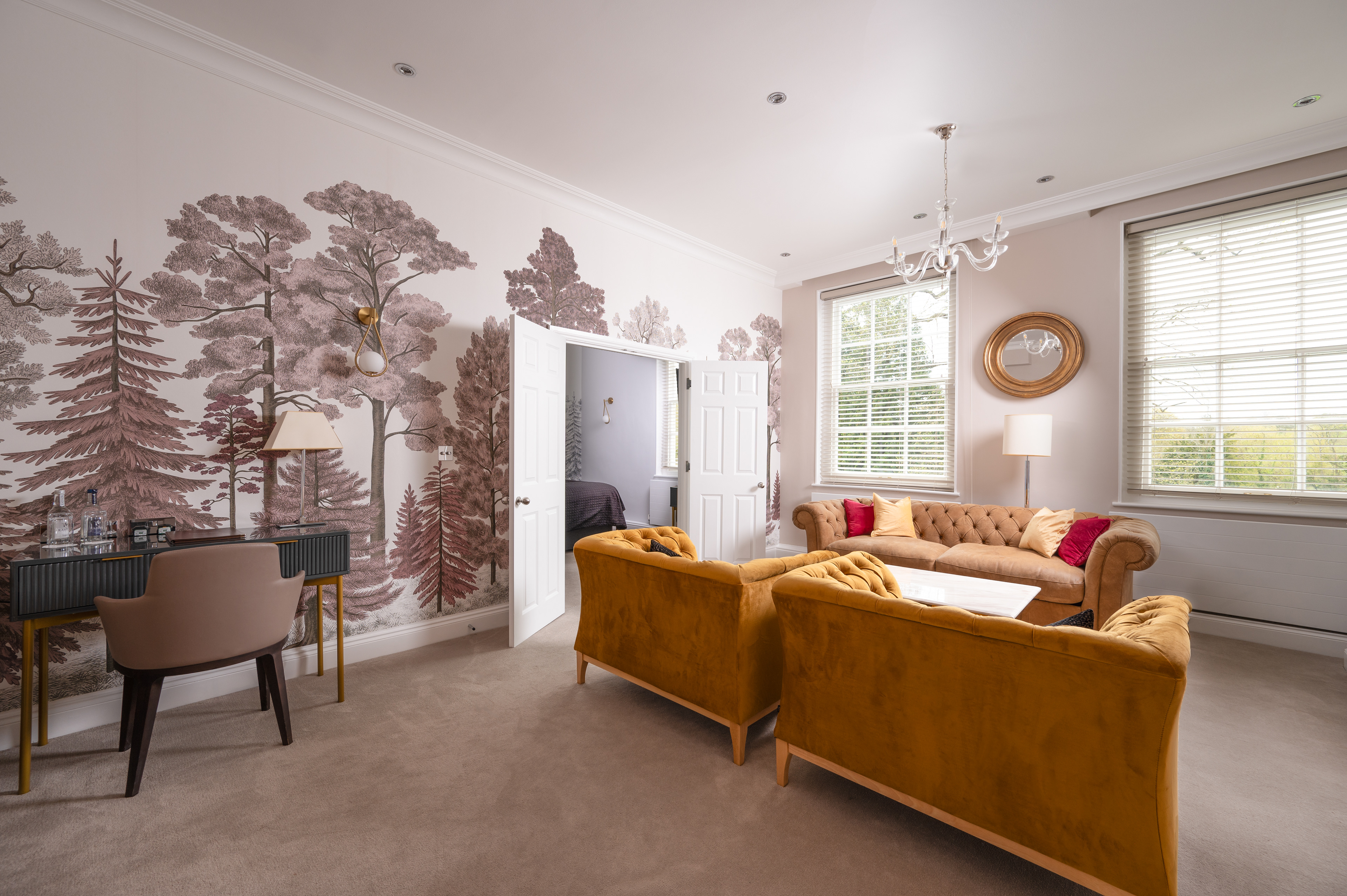 A photograph of the interior of a hotel suite with a writing desk and chair,  yellow velvet arm chairs and sofa. The doors are open to the bedroom showing the corner of the bed in the background.