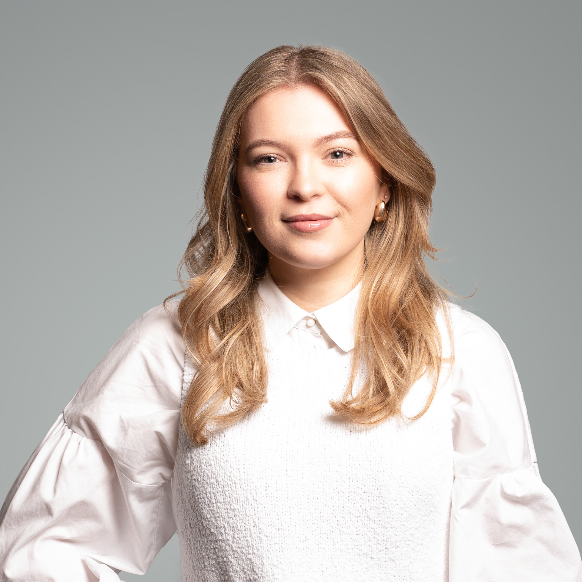 Portrait photograph of a young woman with long blond hair wearing a white shirt and jumper set against a grey background
