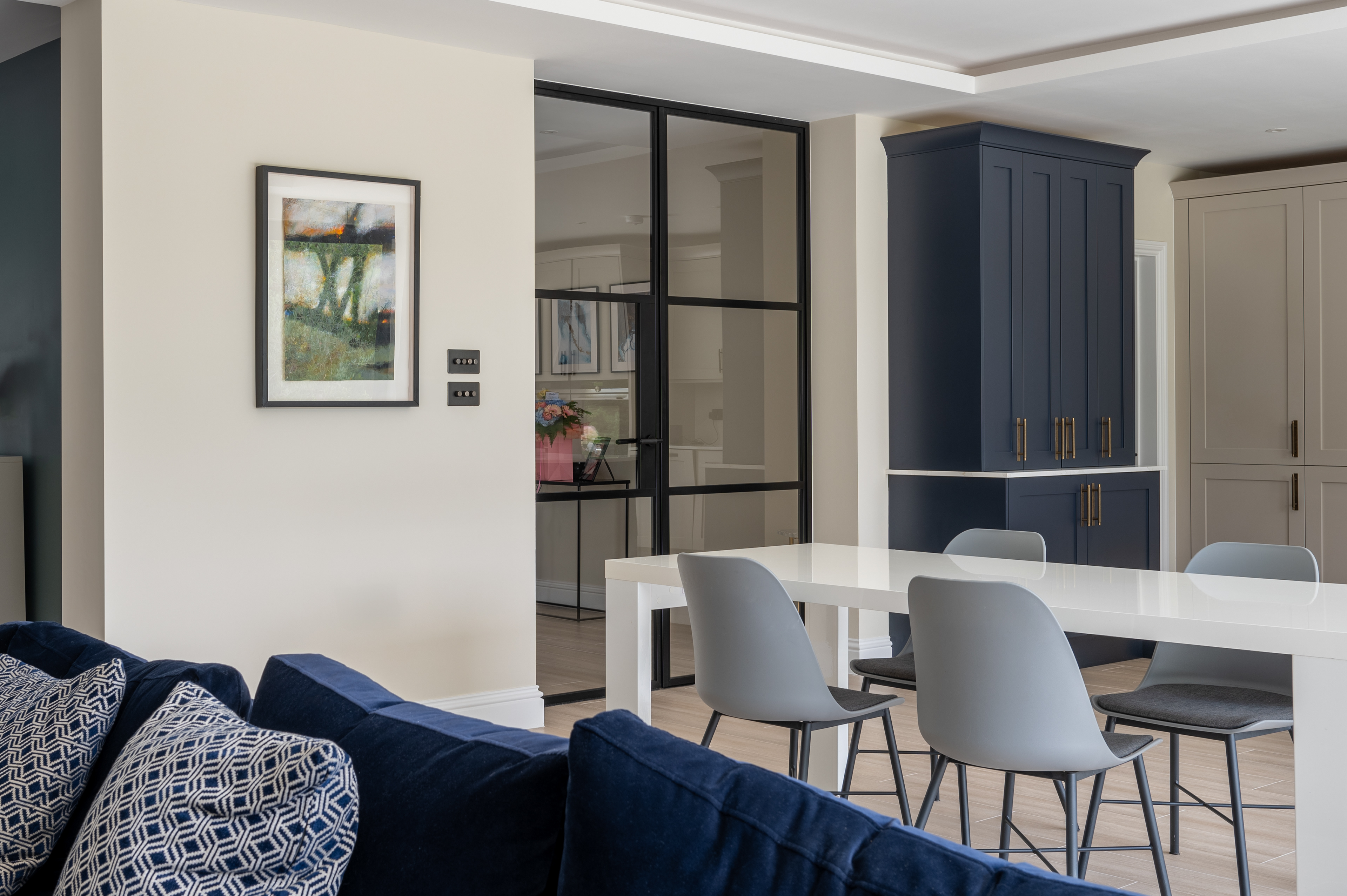 A photograph of the interior of a kitchen, dining and living room. There is a part of a blue sofa in the foreground a dining table and chairs  behind that and kitchen cupboards in the background. The focus of the image is the steel effect industrial style doors that lead to the hallway