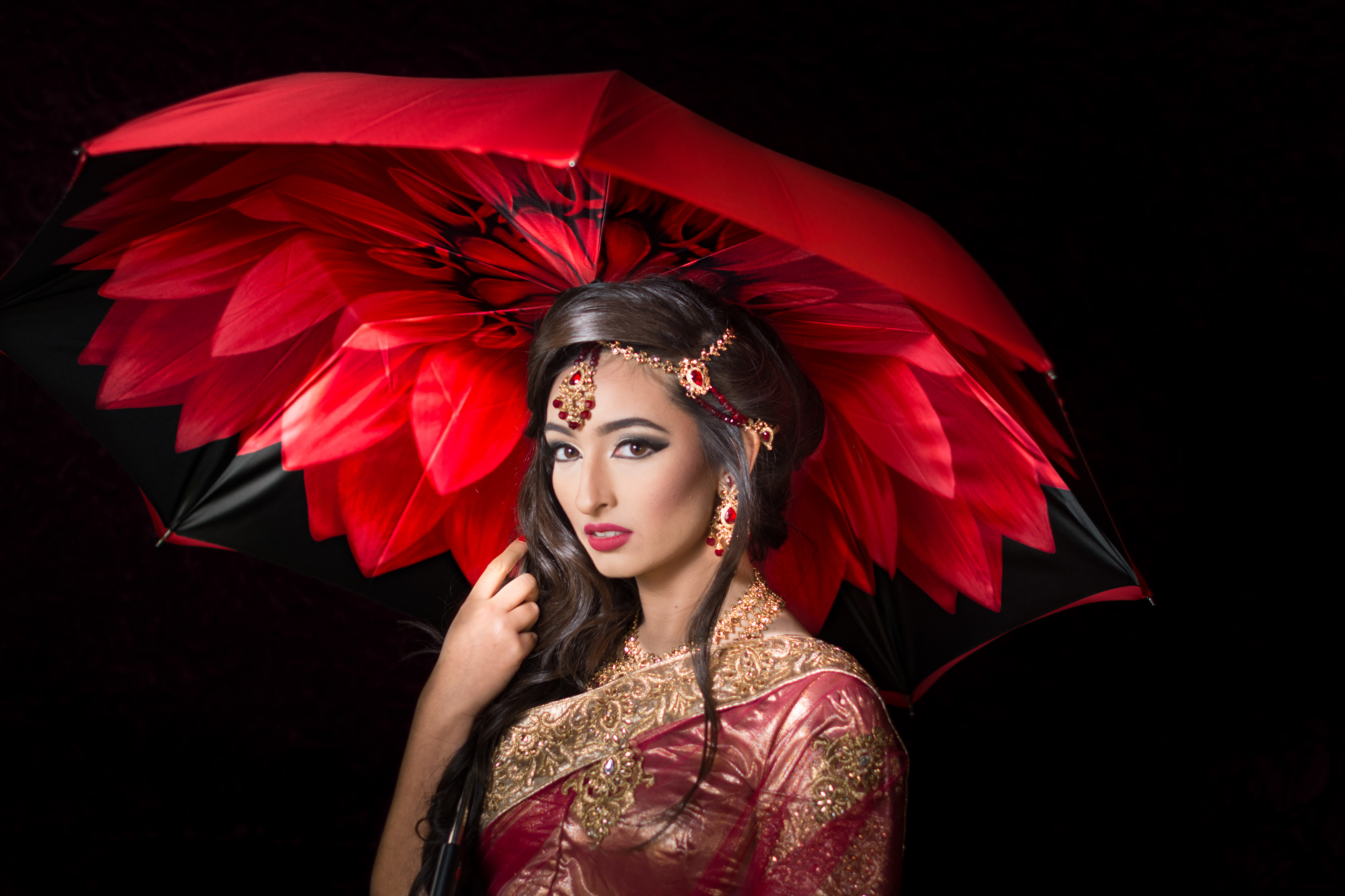 A photograph of a woman wearing a red and gold sari and holding a red and black umbrella