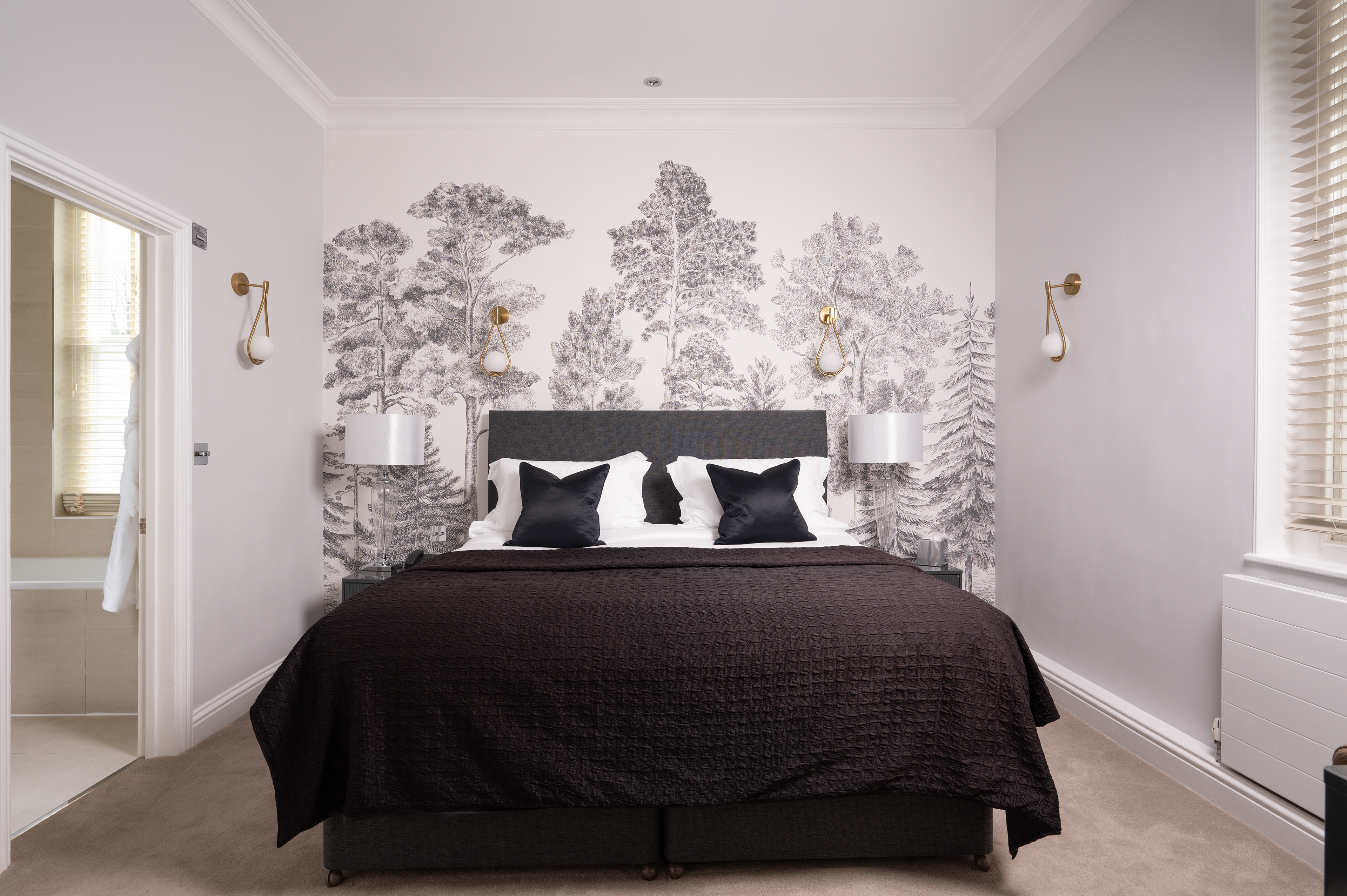 A photograph of a hotel bedroom. The bed has crisp white sheets with black accent cushions and a black throw. The wall behind the bed has wallpaper that is pencil drawings of trees. The en suite bathroom can be seen on the left hand side of the image