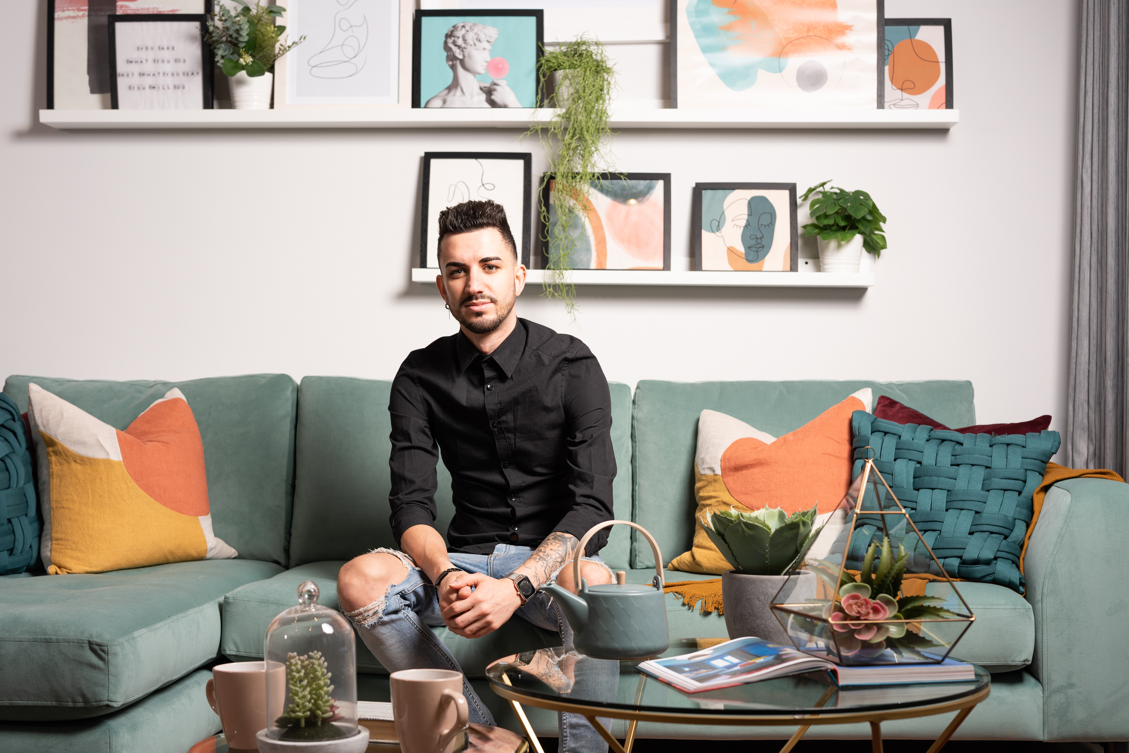 Interior photograph of a young man in ripped jeans and a black shirt sitting on a green L-shaped sofa in a show home