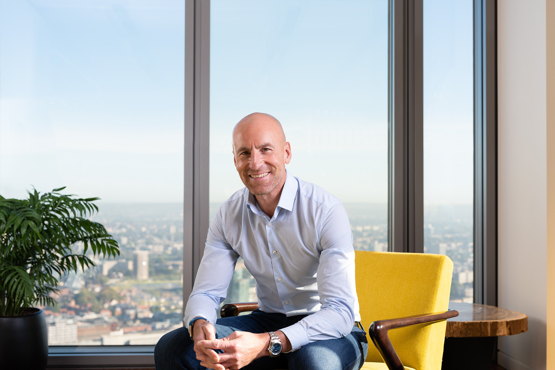 Portrait  photograph of a man sitting in a yellow arm chair. Behind him are views across London.