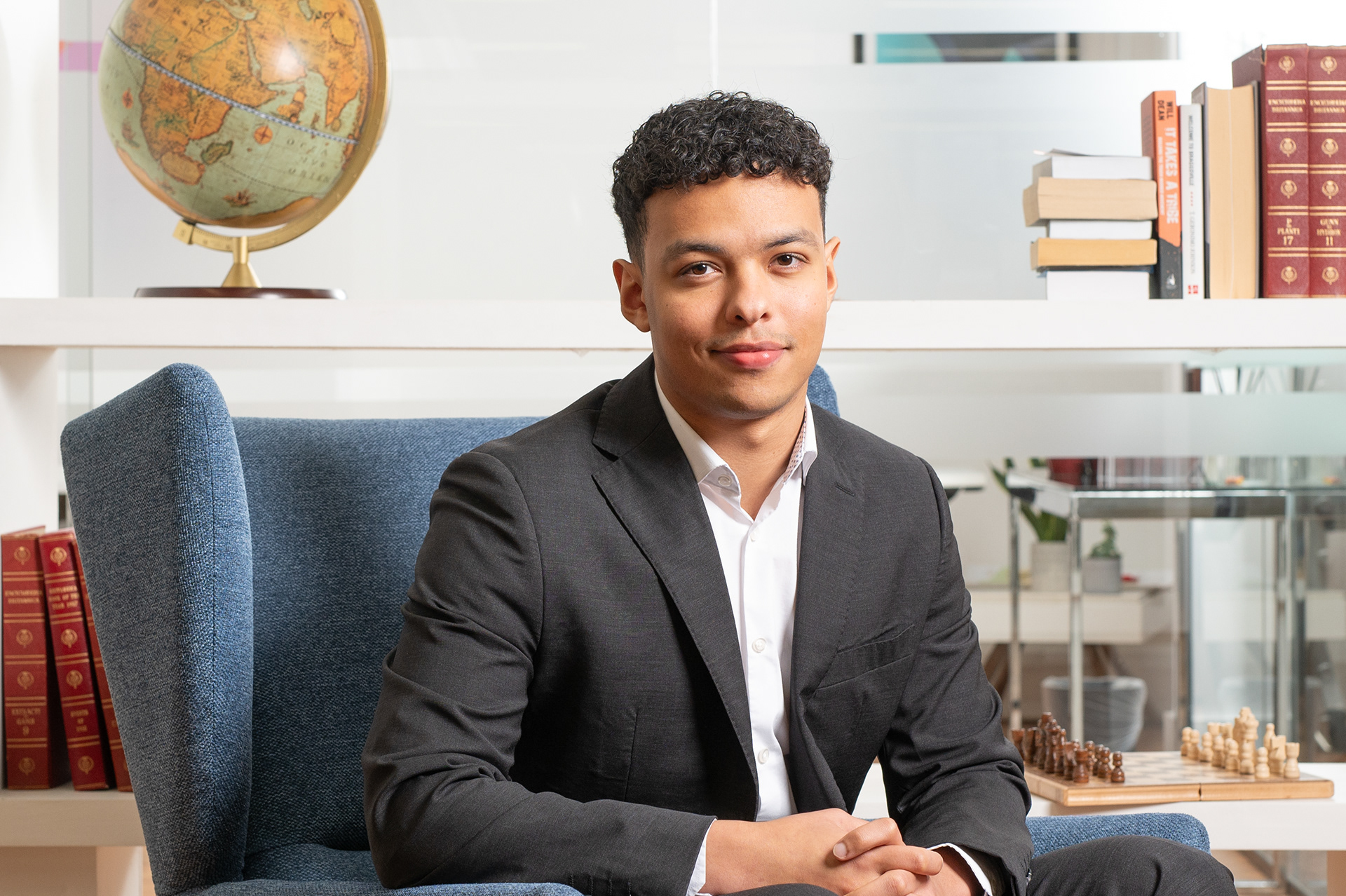 A business headshot of a young man in a suit sat in a blue arm chair