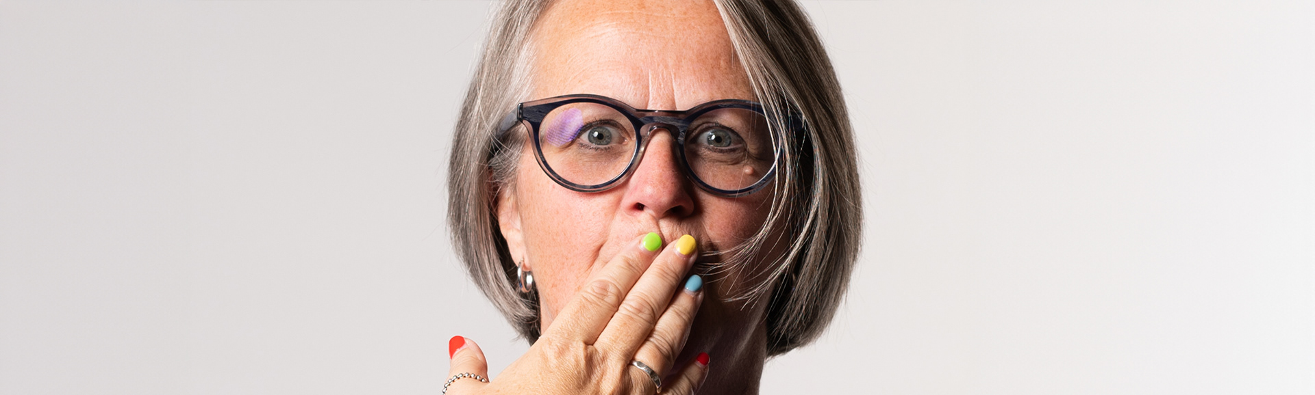 Portrait photograph of a woman with glasses and a bob haircut. Her hand is over her mouth and she has brightly painted finger nails.
