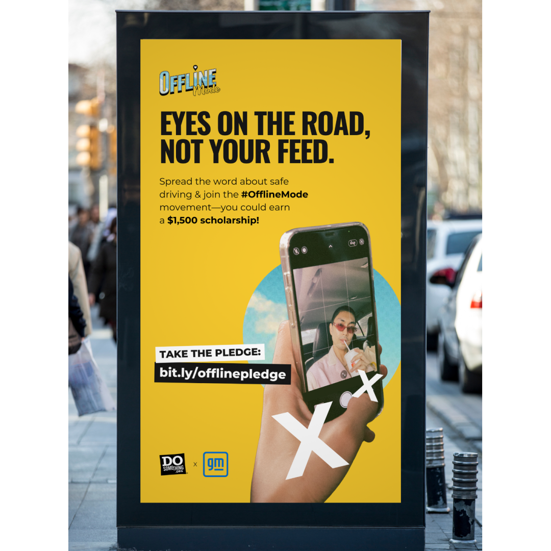Digital billboard for DoSomething and GM’s “Offline Mode” campaign in New York City. The ad features bold text reading “Eyes on the road, not your feed,” alongside a photo of a hand holding a smartphone mid-selfie. A bright yellow background and call-to-action text encourage viewers to take the pledge at a shortlink. Campaign logos are shown at the bottom.