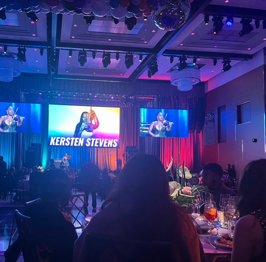 Photo from The Futures Ball gala showcasing violinist Kersten Stevens performing on stage. She is featured on a large screen with rainbow gradient lighting and her name displayed in bold type, flanked by two smaller side screens. The ballroom is lit with deep purples and blues, with chandeliers, a disco ball, and elegant table settings visible in the foreground. Guests are seated at round tables, creating a vibrant, immersive atmosphere that blends performance, celebration, and youth-centered design. Captures the live production quality and emotional tone of the gala experience.