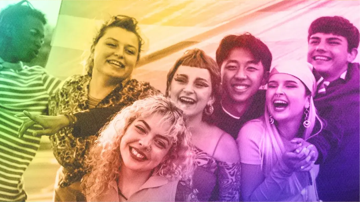A diverse group of seven young people stands together, smiling and laughing in a joyful, candid moment. They are framed by a soft rainbow gradient overlay, symbolizing LGBTQ+ pride. The group includes individuals of varied racial backgrounds, genders, and styles, reflecting youth self-expression and inclusivity. The accompanying text notes that the photography was chosen to highlight energy, diversity, and representation in the context of conversations around banned books.