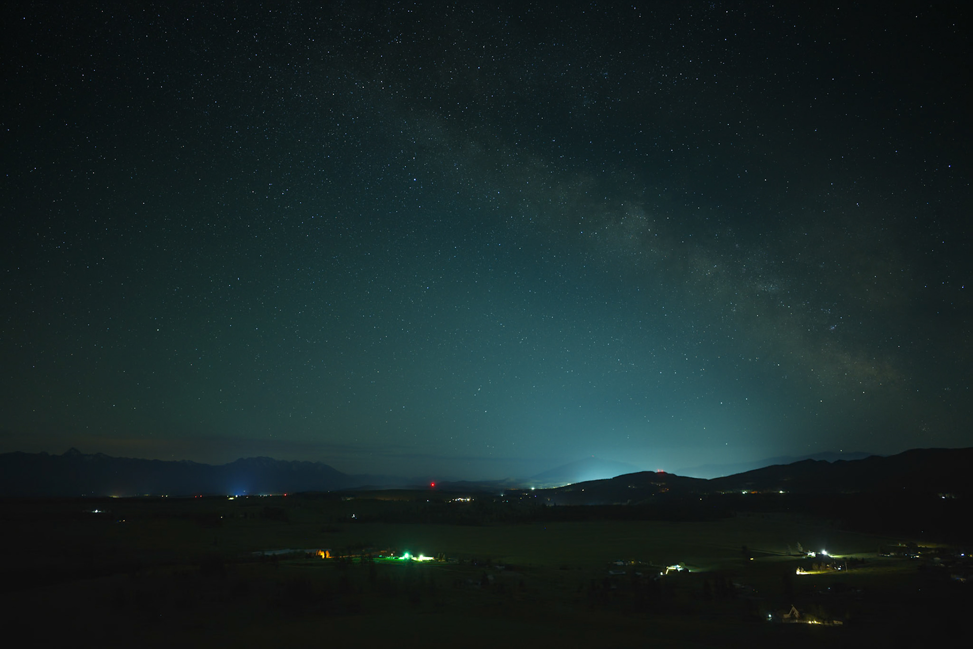 Looking south from Wycliffe Buttes near Cranbrook, British Columbia, the Milky Way spans the night sky above ranchlands and distant mountain ranges. Light from Cranbrook glows faintly on the horizon, highlighting the contrast between the dark sky and the settled landscape below.