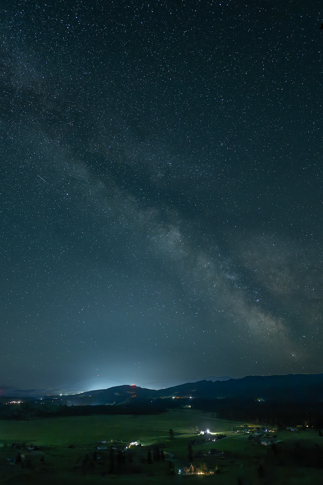 This vertical nightscape captures the Milky Way arching south over ranchlands and mountain foothills, as seen from Wycliffe Buttes near Cranbrook, British Columbia.