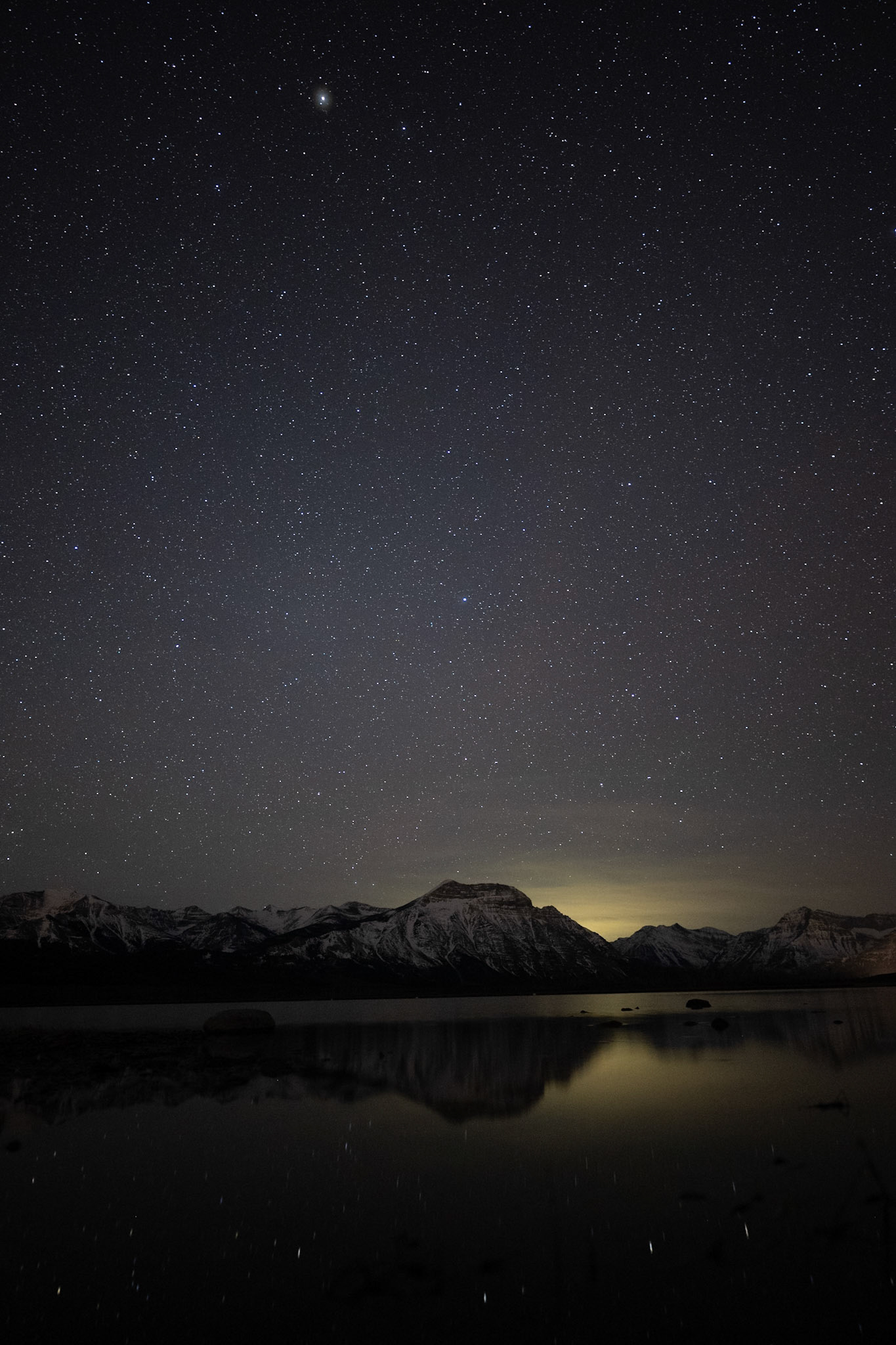 The Milky Way rises above Waterton Lakes, reflecting in still waters below. Faint town light silhouettes the mountain skyline on a spring night.