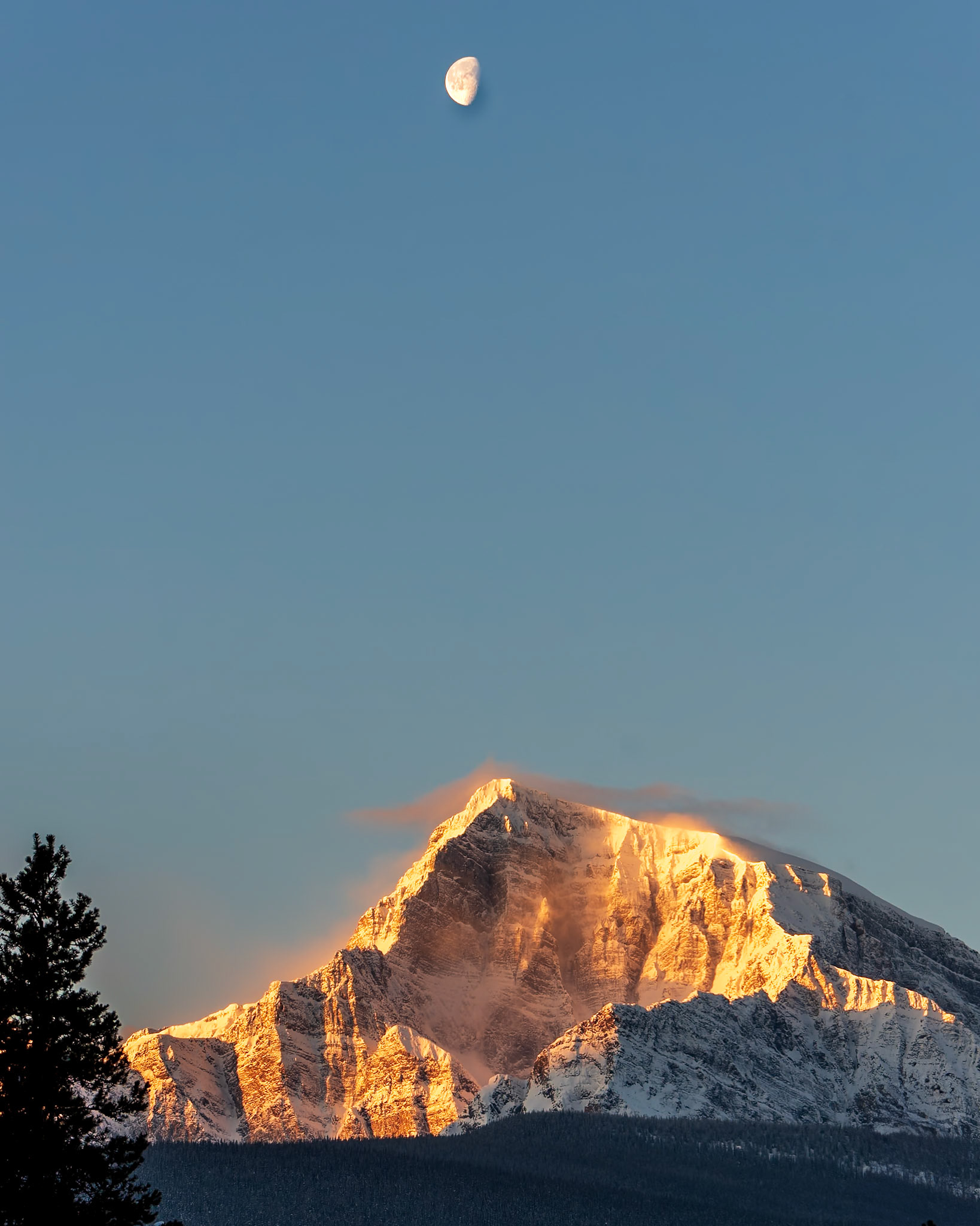 The moon rises over an isolated peak in the mountains of Banff National Park.