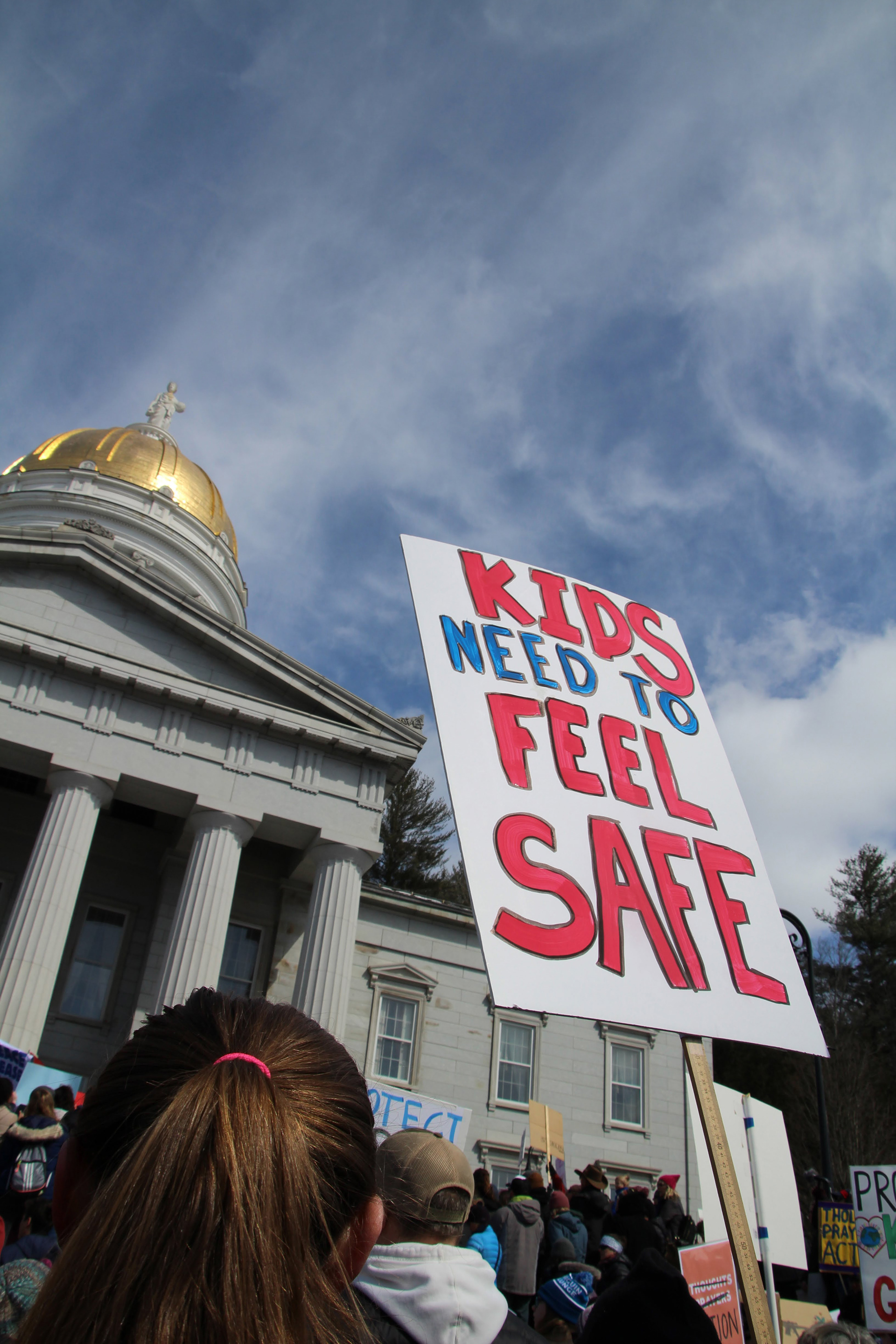 A sign is held by the golden dome of the Vermont State Capitol in Montpelier, Vt. on March 24, 2018. (Lia Chien)