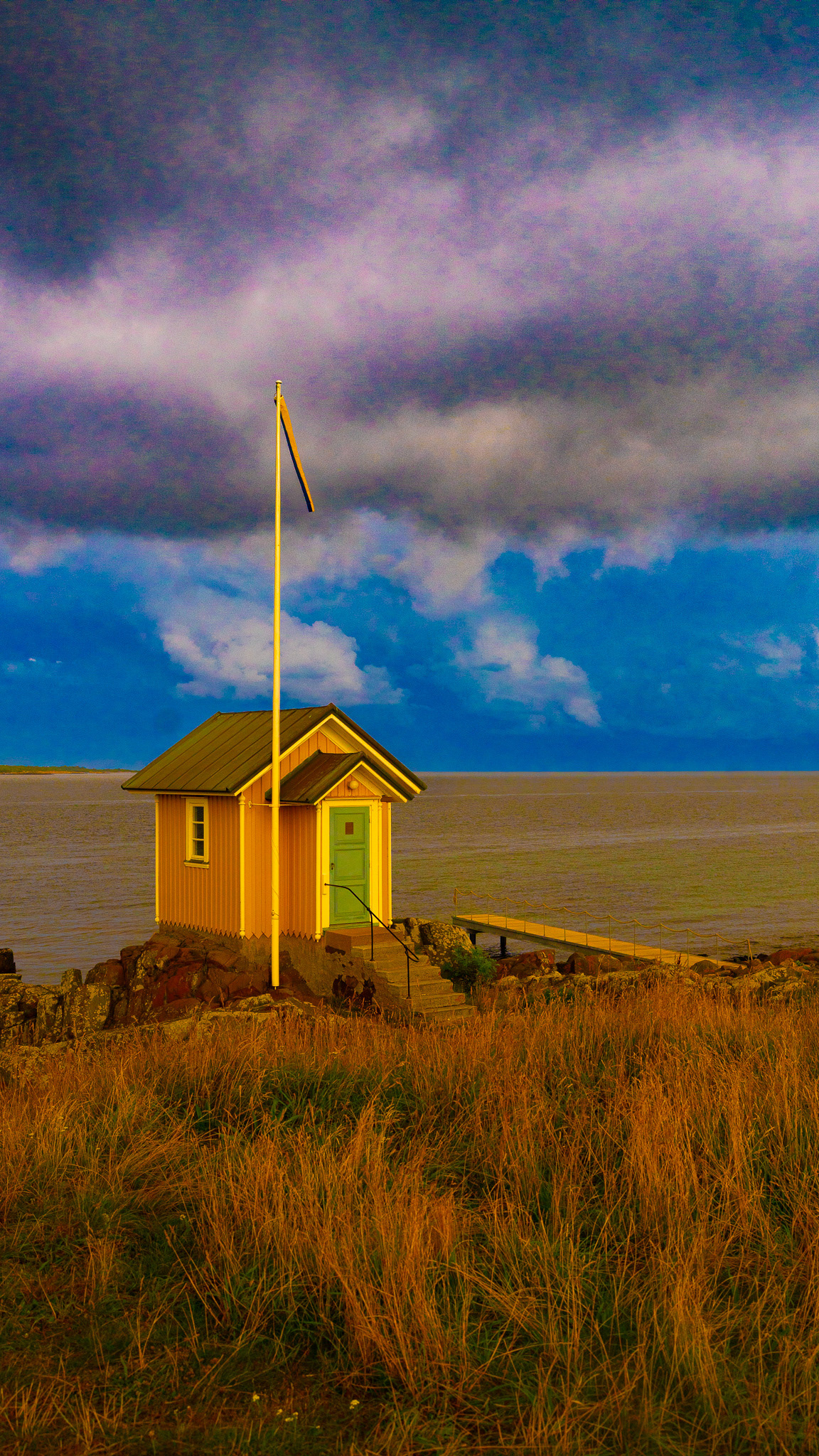 A small yellow seaside cabin with a green door stands on a rocky outcrop in Torekov, Sweden, beside a calm sea. A tall flagpole with a Swedish pennant rises beside the cabin, and a narrow jetty stretches out into the water. The sky is dramatic, with dark, stormy clouds contrasting against a vibrant, golden foreground of dry grass.