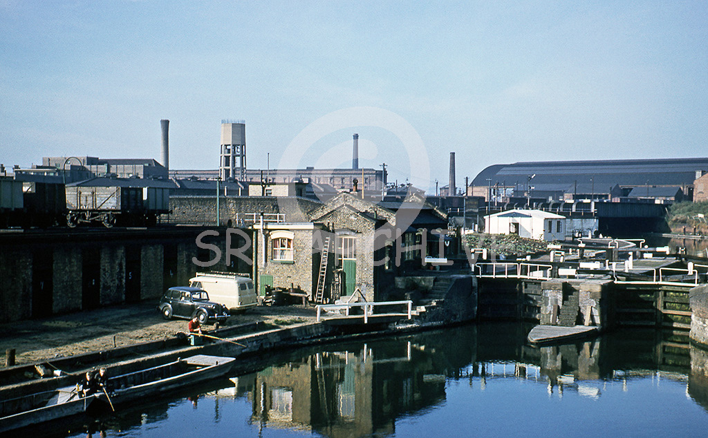 Regent's Canal Beyond St Pancras in May 1959. Grade 11 lock keepers cottage with a fisherman on the river bank. In the background we see the concrete water tower and chimney marking the site of the St Pancras Hospital. Also further back we see the MR Granary Warehouse and ale store with the signals identifying the course of the Midland main line. SRL No 37   