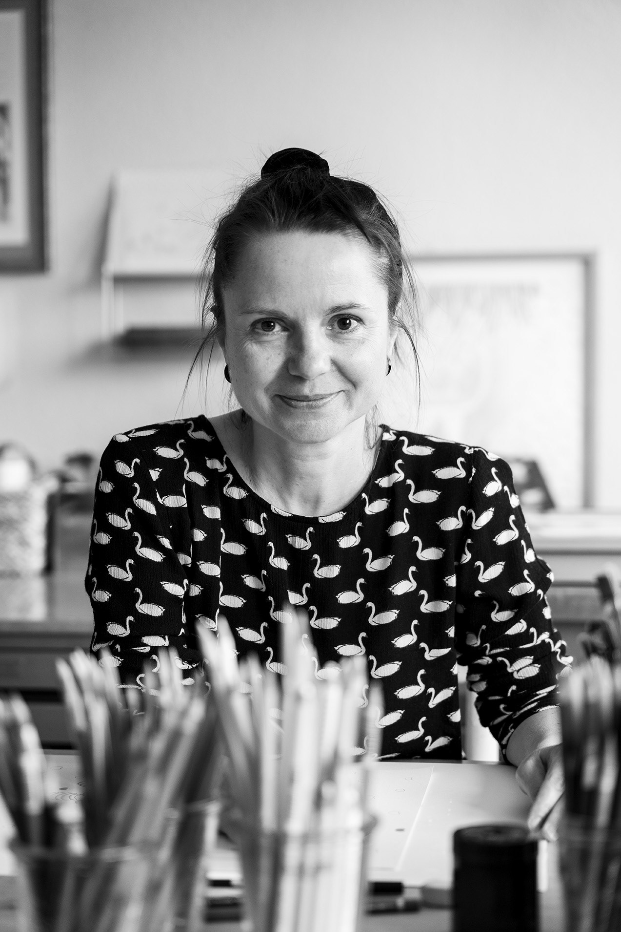 Black and white portrait of the artist Nina Midi, taken in her atelier. Artist is sitting at the desk with pencils in foreground