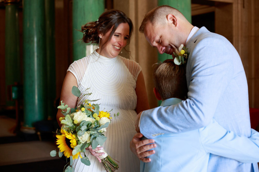 Groom comforts his son in hallway as bride smiles at them both