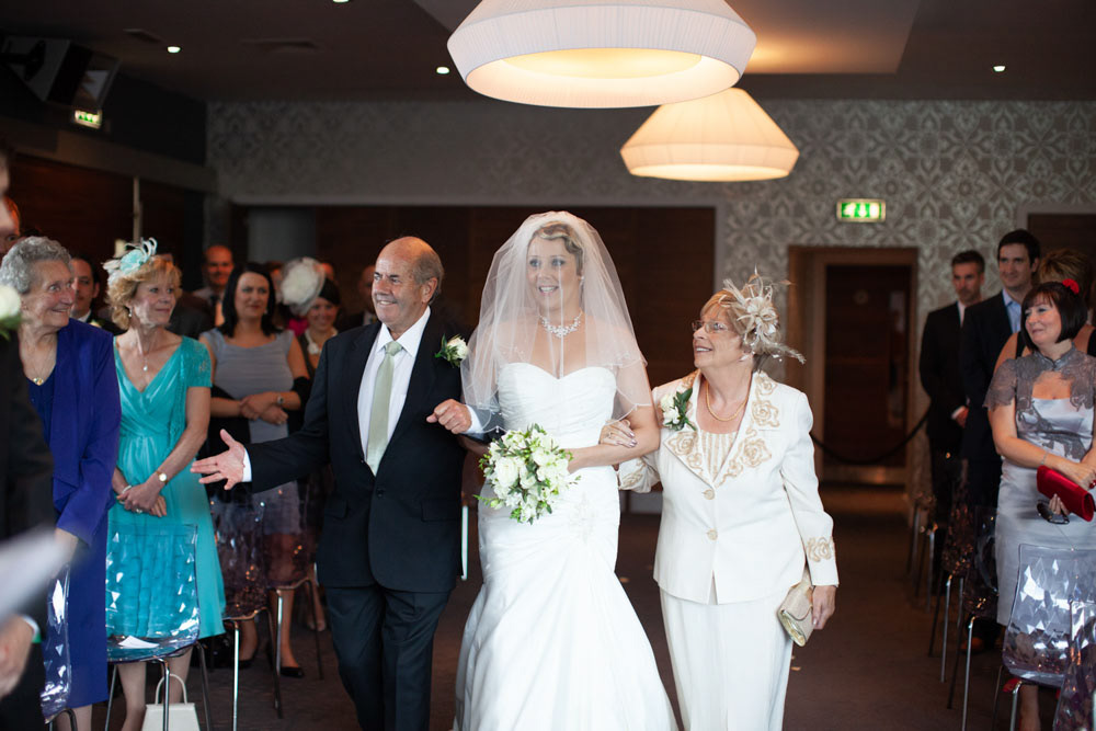 Bride walks in with her parents, one on each arm, beaming with pride.