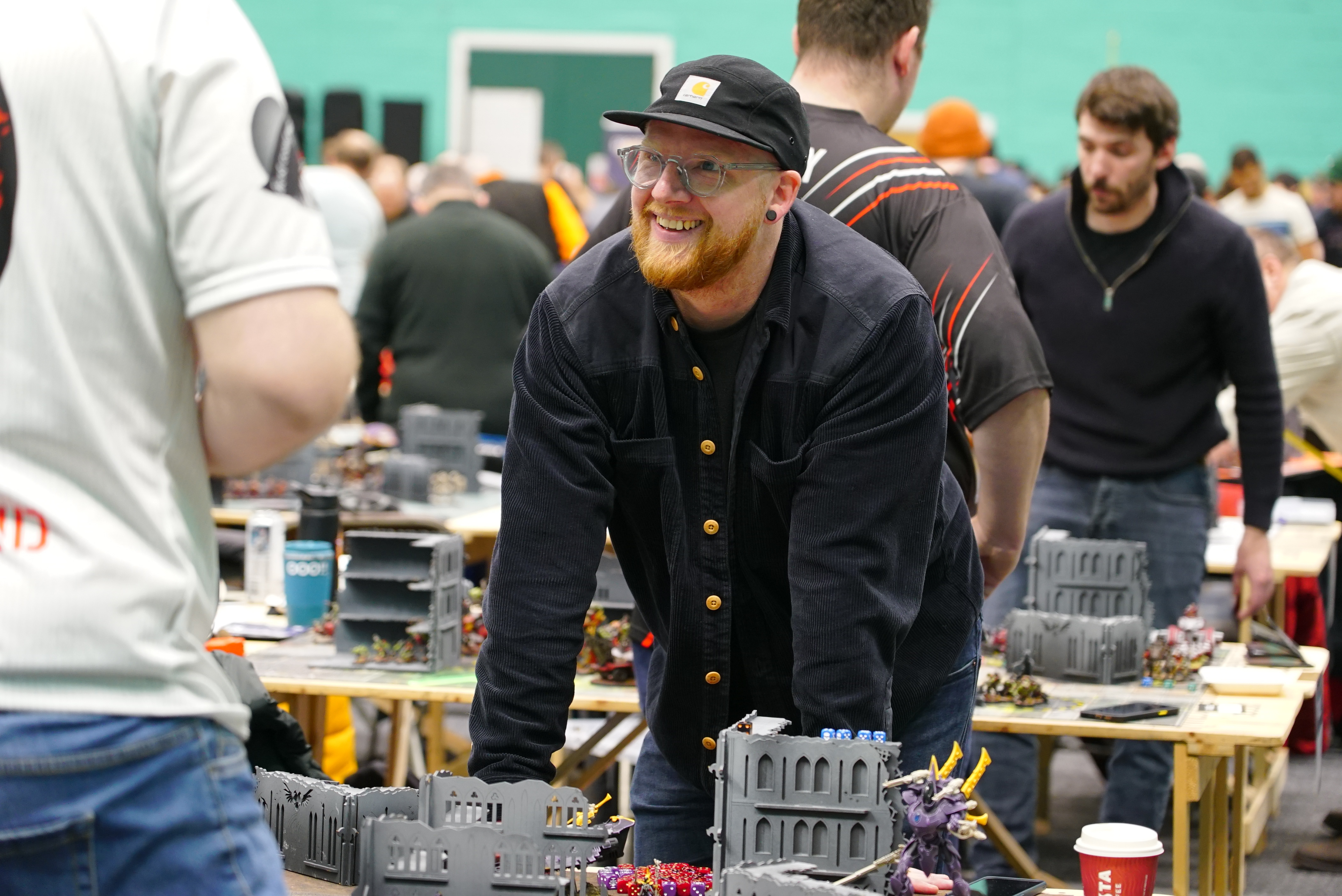 a man in a hat and glasses smiling at a table with small buildings and warhammer 40k models.